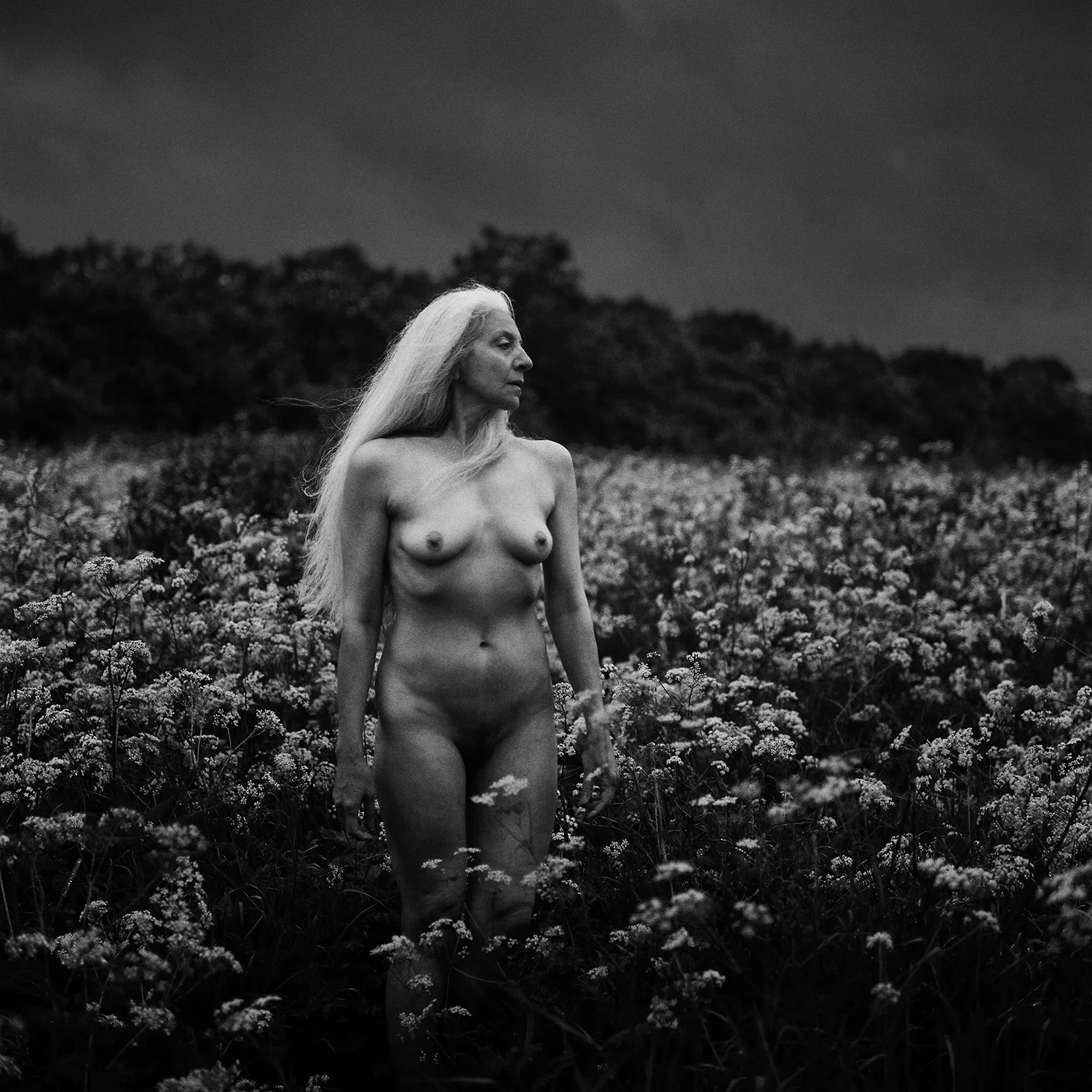 Black and white photo of an older nude woman with long hair standing in a field of flowers, looking to the left.