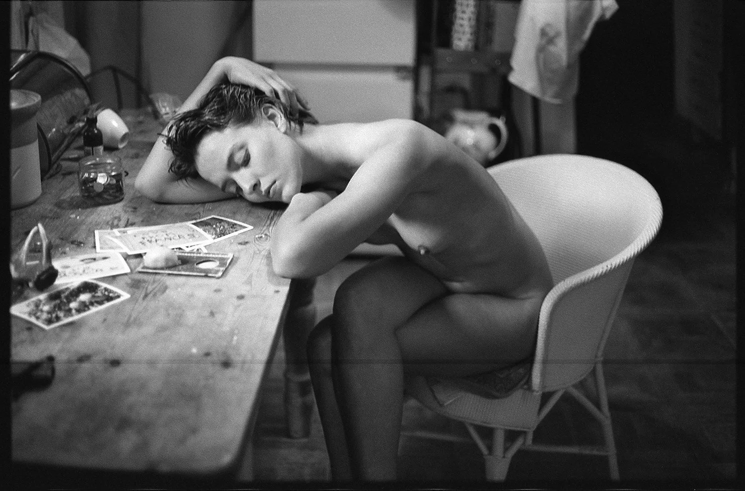 A woman with short hair and makeup, sitting nude in a chair with her eyes closed, resting her head on her crossed arms on the table covered with photographs, jewelry, and personal items.