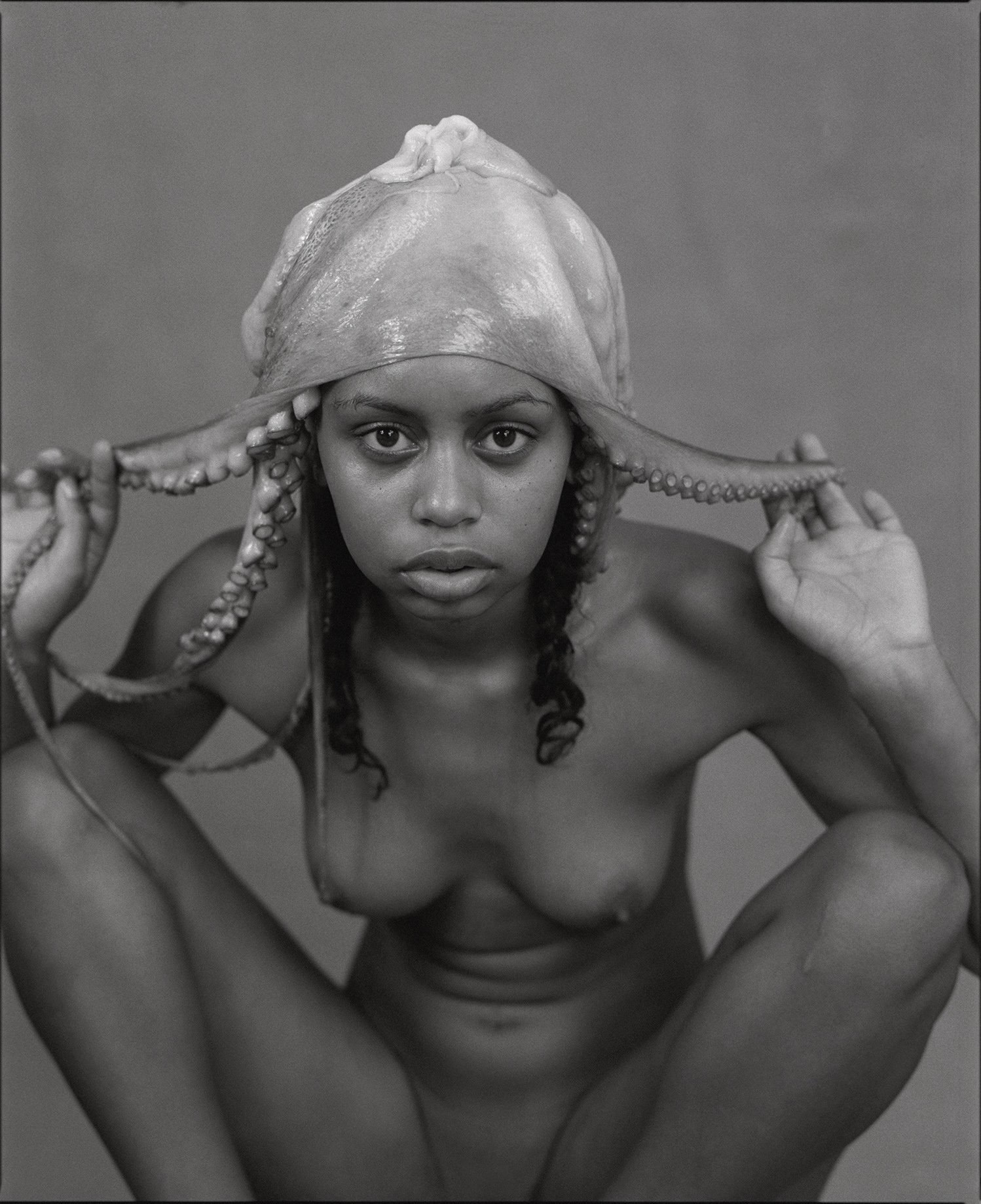 Black and white photograph of a nude woman with curly hair, squatting and looking directly at the camera, wearing a large octopus hat.