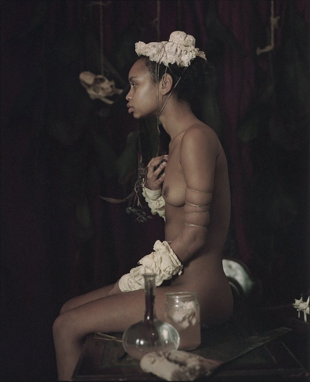 A nude woman sitting sideways on a wooden surface, wearing a large, ruffled headpiece and arm accessories. She is in a dimly lit room with dark background, surrounded by glass bottles and jars.