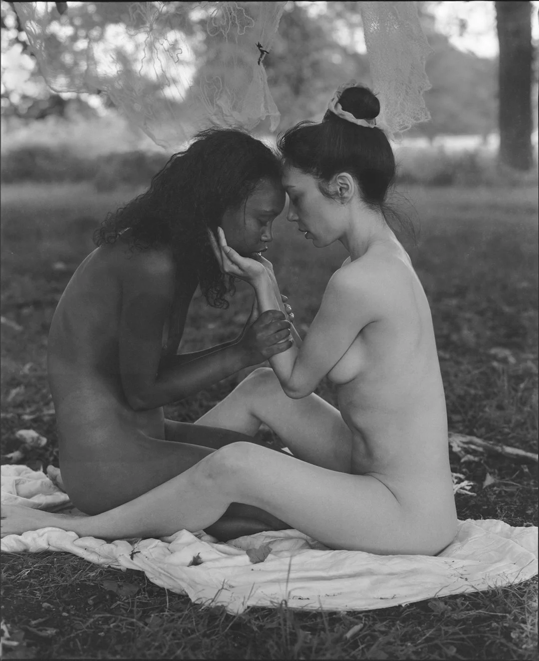 Two women, one with dark curly hair and the other with dark straight hair in a bun, sit intimately on a blanket in a natural outdoor setting, touching foreheads with closed eyes.