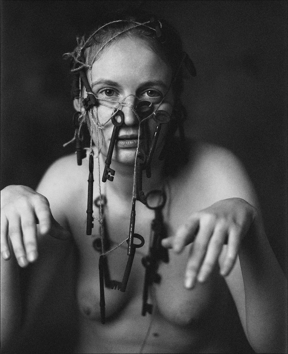 Black and white photo of a topless woman with dreadlocks, face, and upper body visible, with various keys hanging on her face and neck, looking directly at the camera.