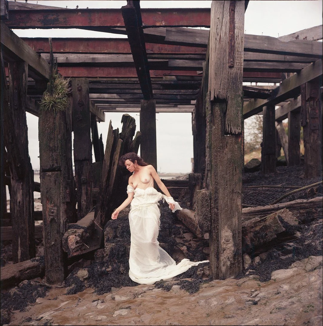 A woman in a white dress standing under a decaying wooden pier on a sandy beach.