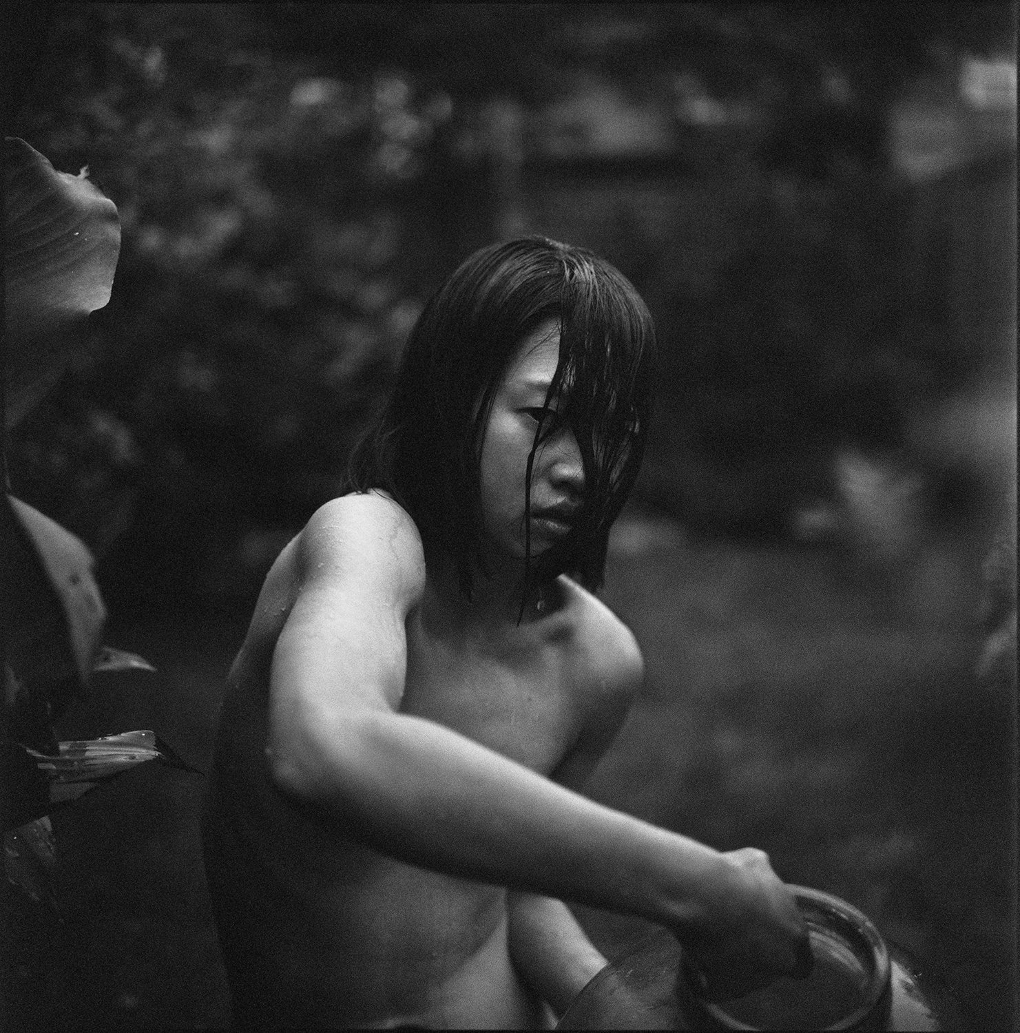 Black and white photo of a young woman with wet hair, sitting partially nude with her arm extended, and looking downward with a serious expression.