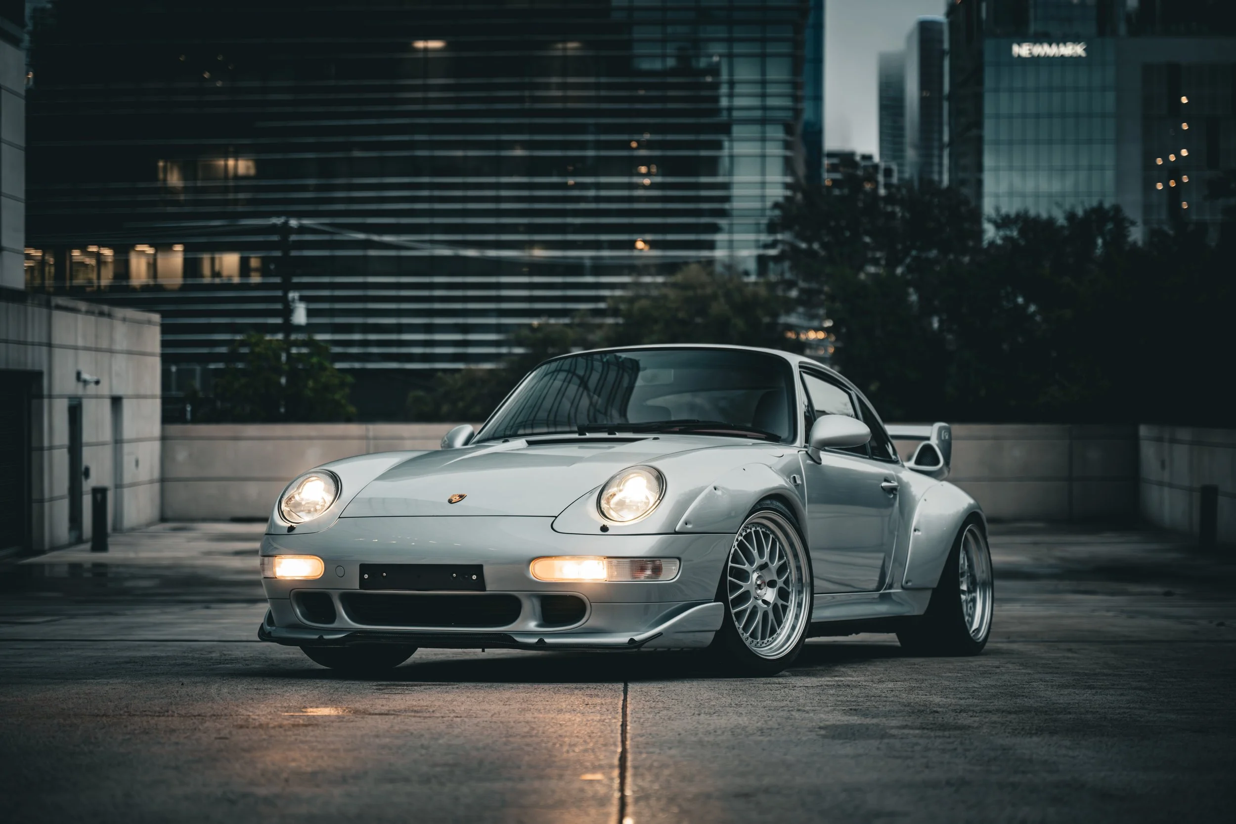 A silver Porsche 993 Turbo GT2 Tribute 911 sports car parked on an urban rooftop parking lot with modern glass buildings in the background during twilight.