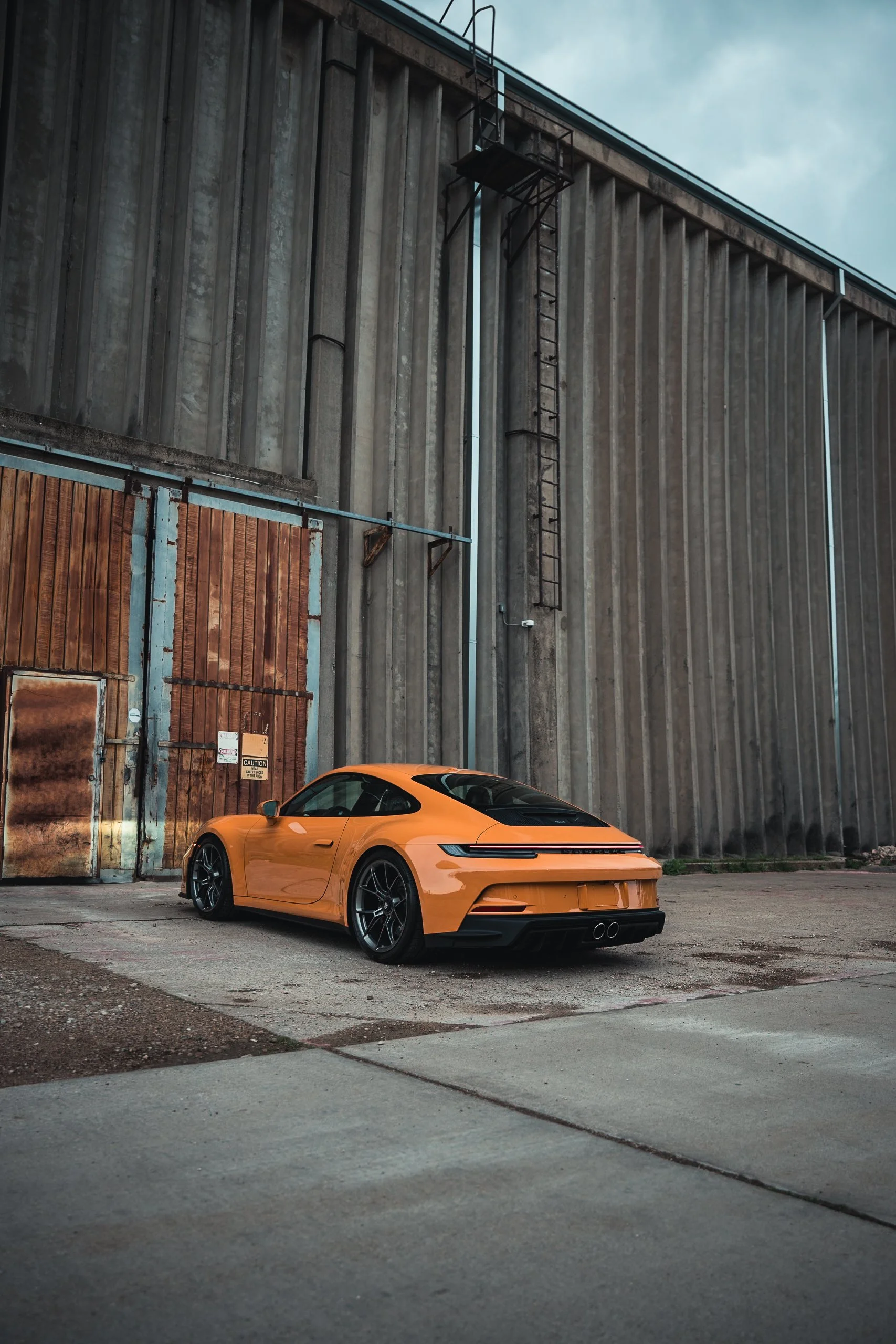 Bahama Orange Porsche 911 GT3 sports car parked in front of a weathered industrial building with metal and wooden door, against a cloudy sky.