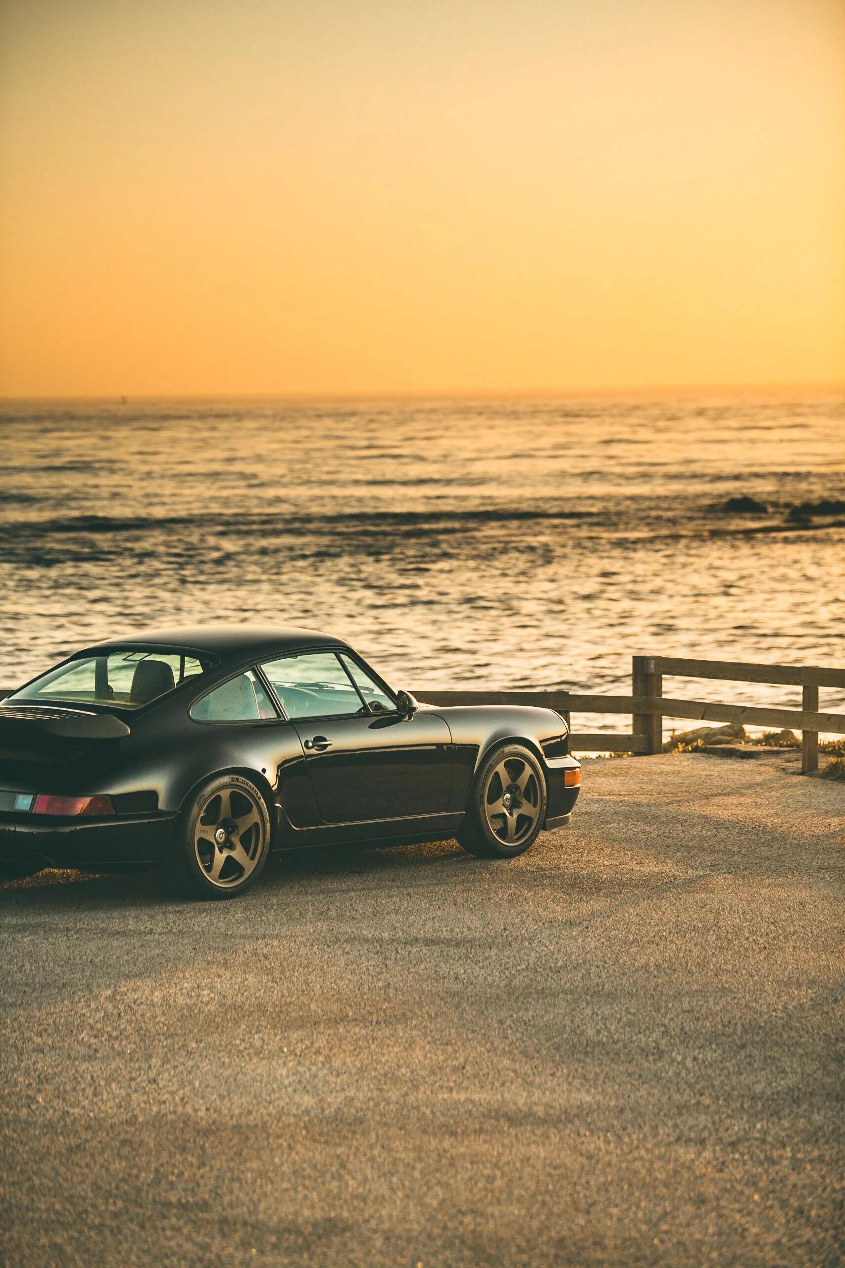 Black Porsche car parked near ocean at sunset with waves and a wooden barrier in the background.