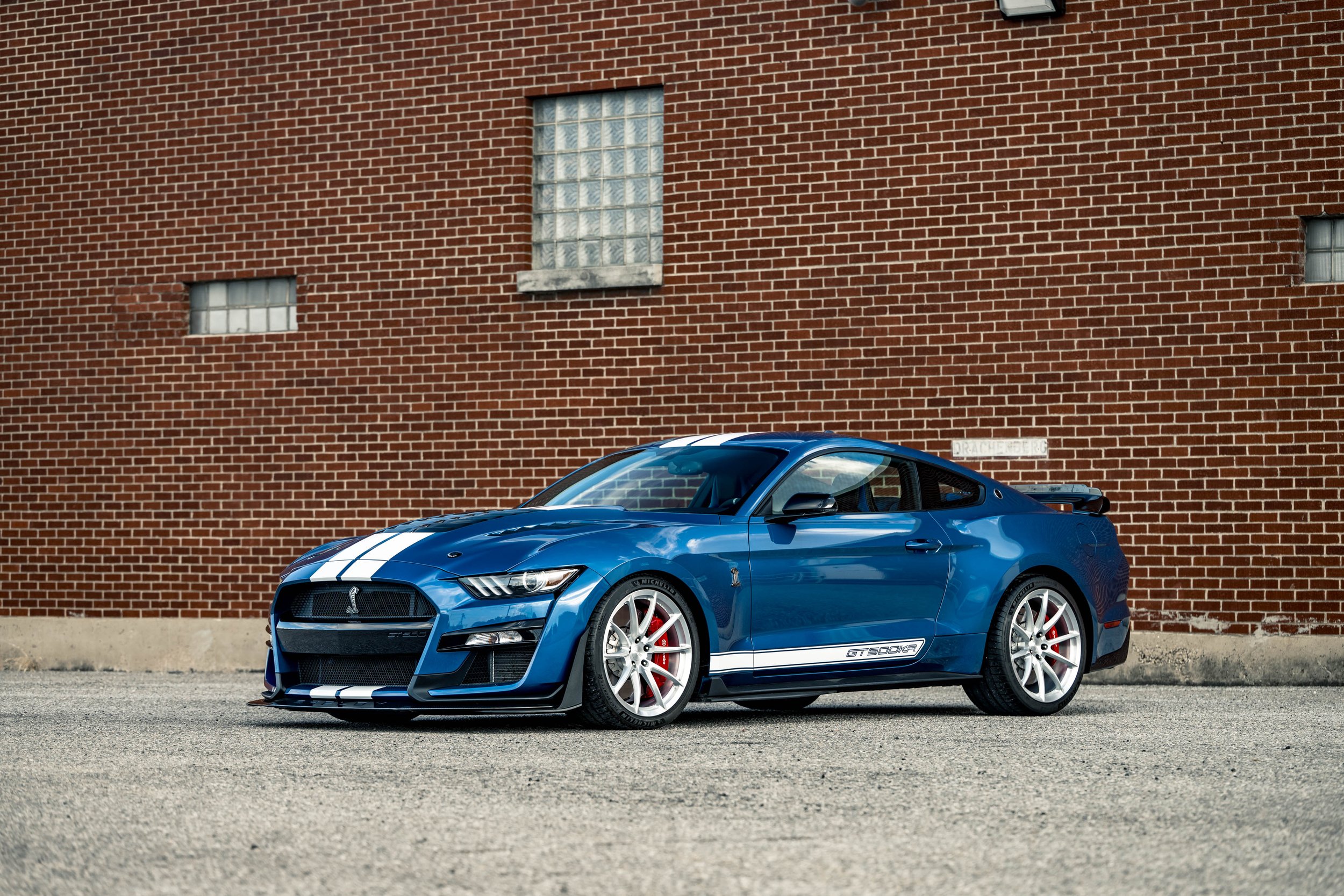 Blue Ford Mustang Shelby GT500 car with racing stripes parked in front of a brick wall.