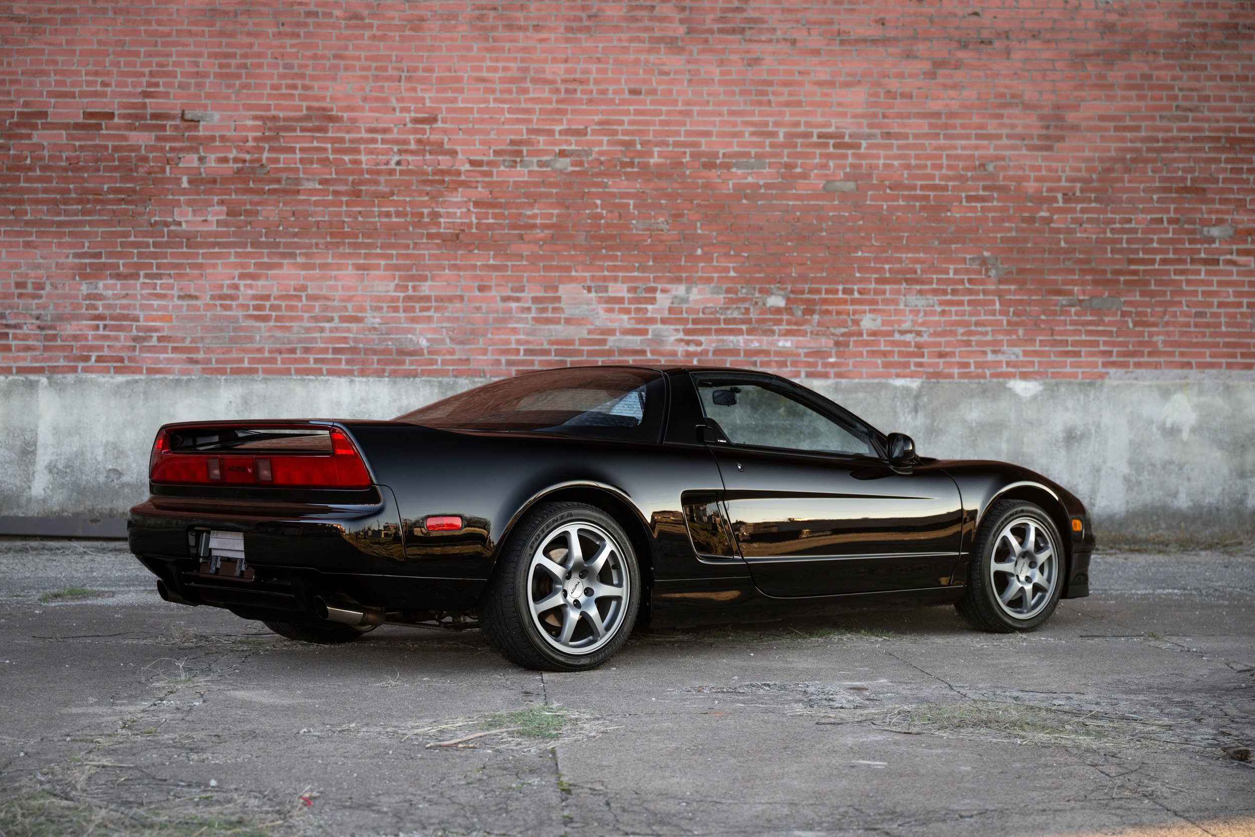 A black sports car parked on a concrete ground in front of a red brick wall.