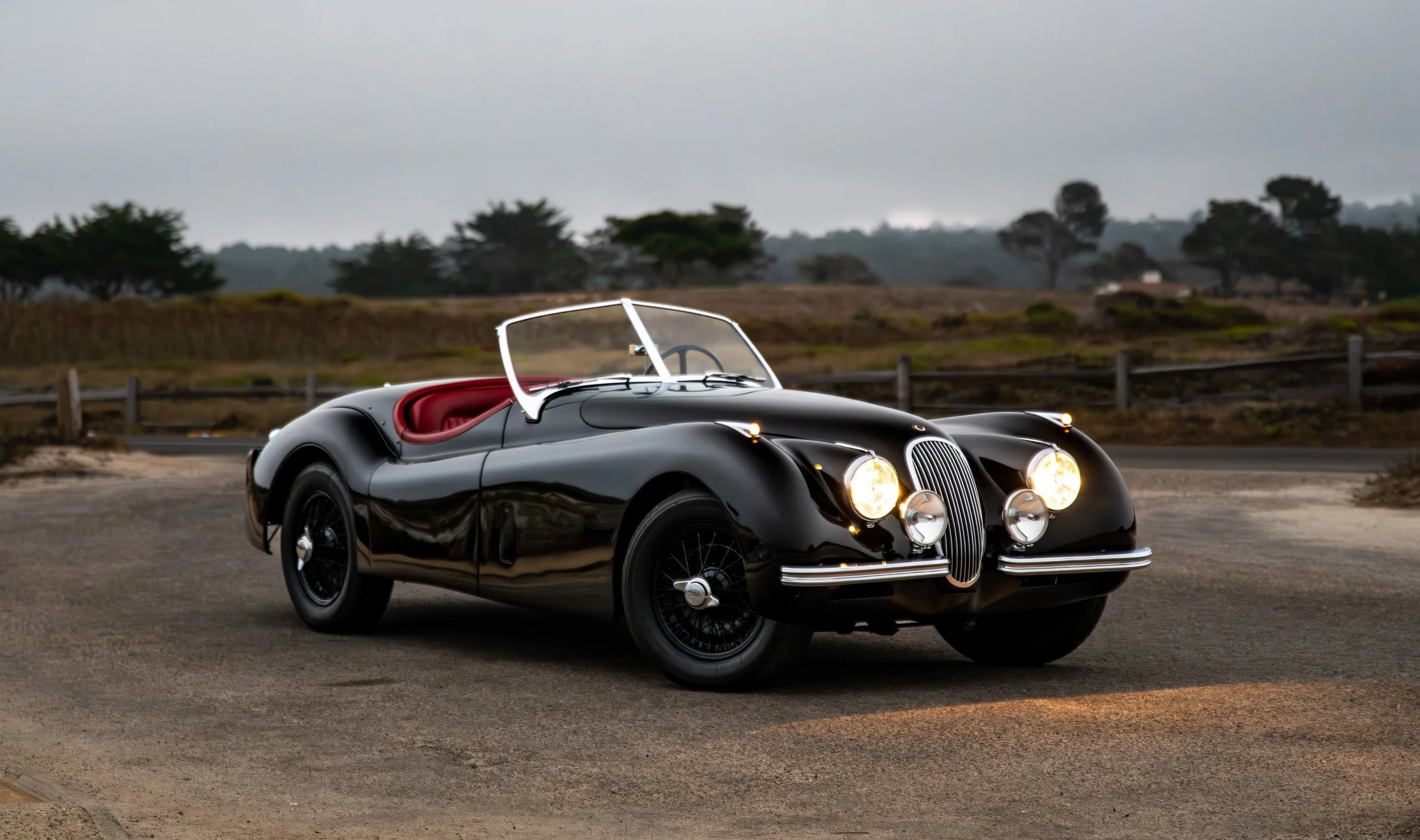 A vintage black convertible car with red interior parked on a road with a rural landscape in the background.