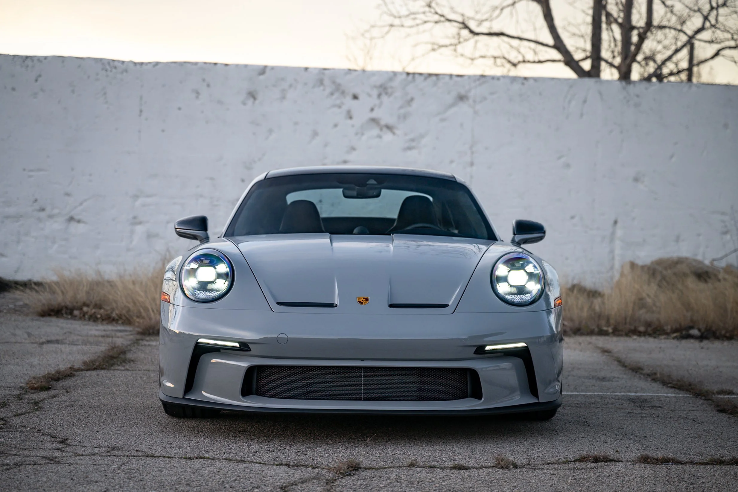 Gray Porsche sports car parked outdoors in front of a white wall with trees in the background.