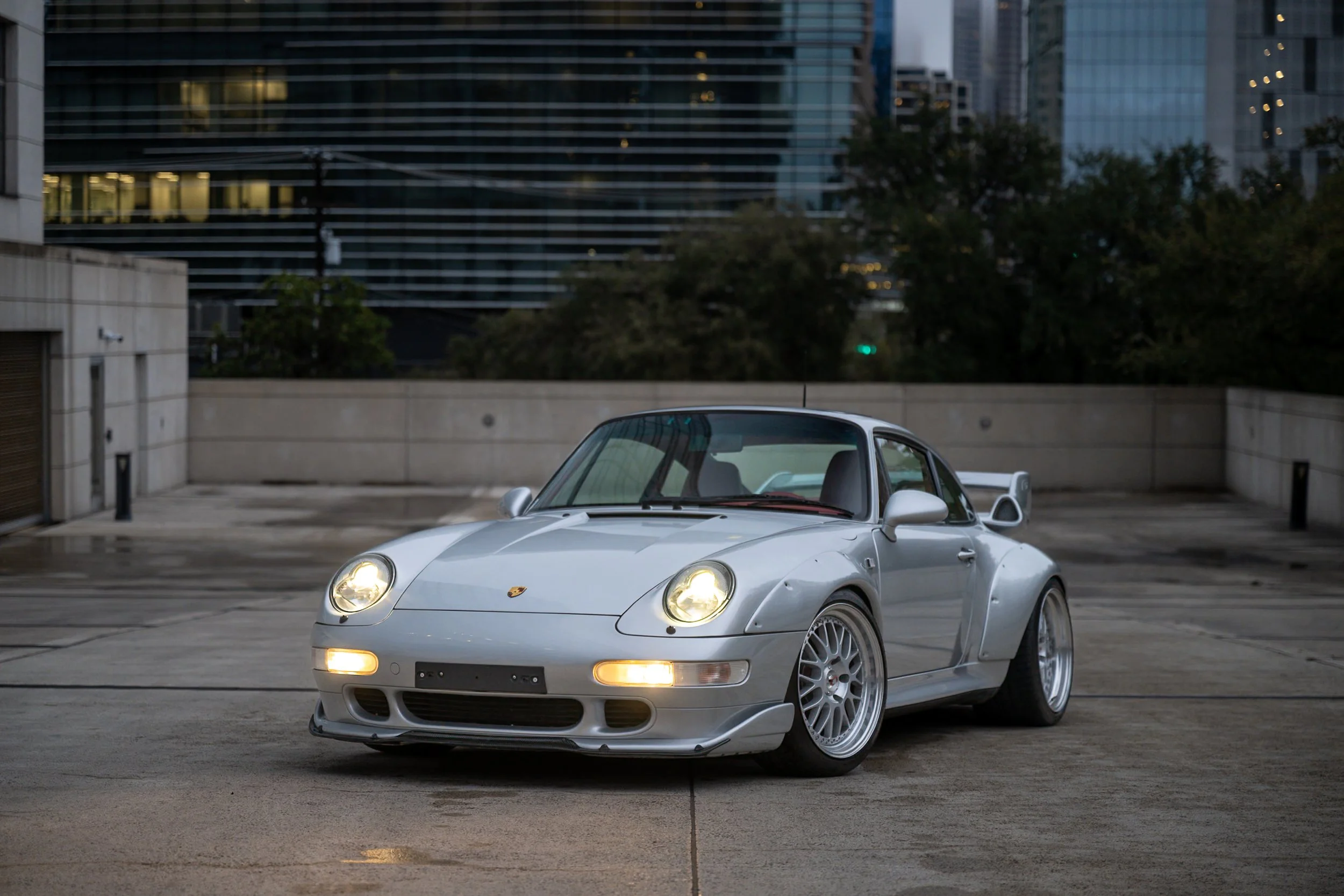 Silver Porsche 911 sports car parked in an urban parking lot at dusk with modern buildings in the background.