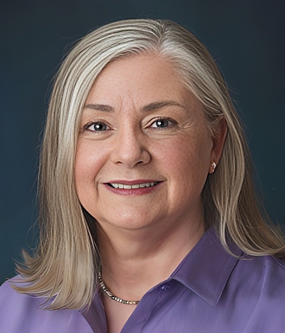 Portrait of a middle-aged woman with blonde hair, blue eyes, wearing a purple blouse, earring, and a necklace, smiling against a dark background.