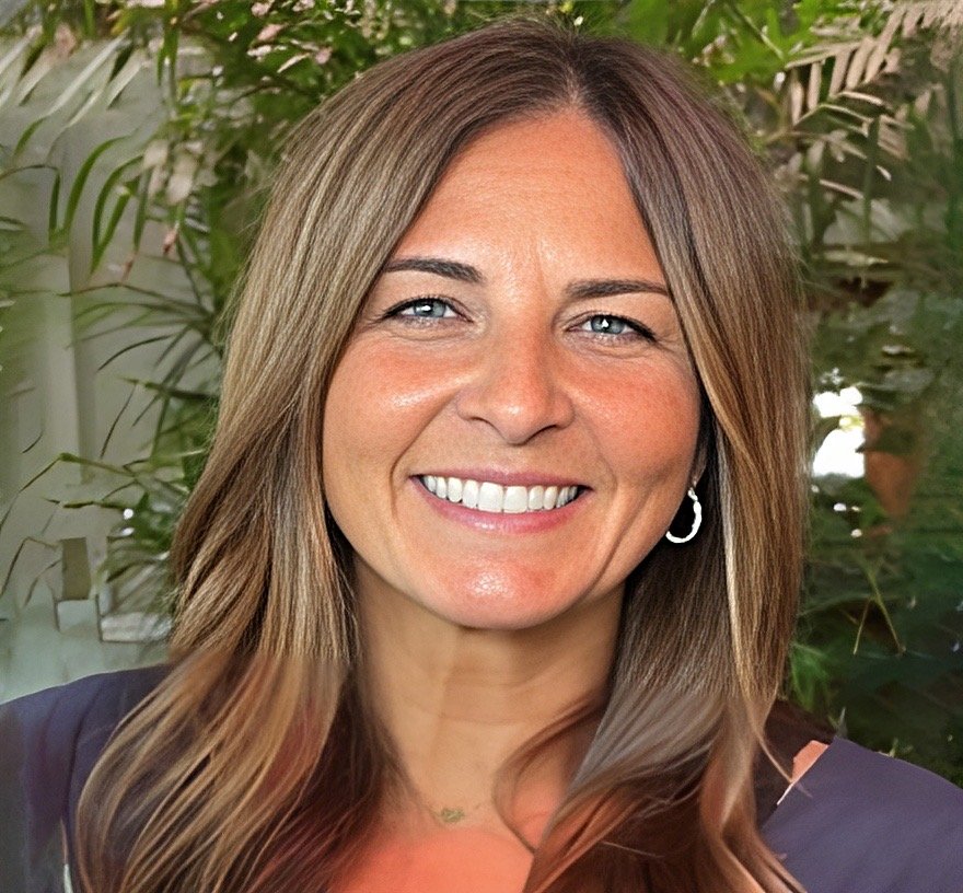 A woman with long, wavy brown hair and blue eyes smiling, standing in front of green foliage.