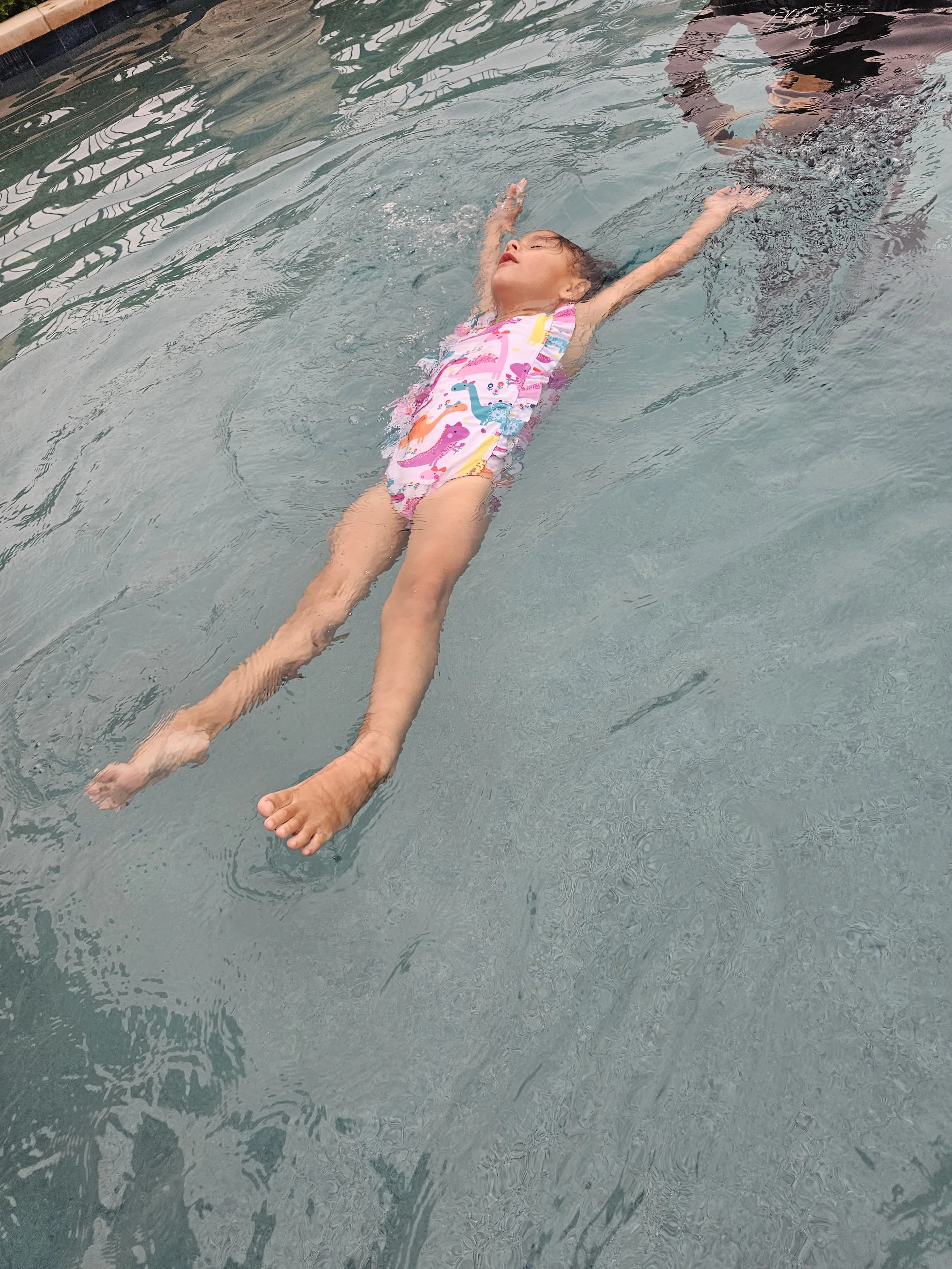 A young girl in a colorful swimwear floating on her back in a swimming pool with her arms extended outward and eyes closed, enjoying the water.