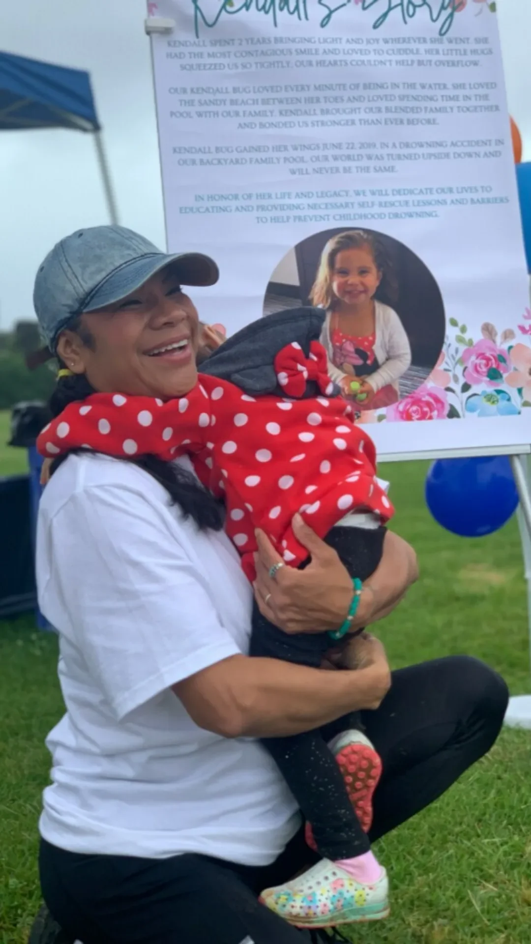 A woman wearing a gray cap, white t-shirt, and a red polka-dot scarf is kneeling on the grass, holding a young girl dressed in black with colorful shoes and a red polka-dot scarf. The woman is smiling and holding the girl close. Behind them, there is a poster with a photo of the girl and some text, outdoors with a blue tent and grass visible.