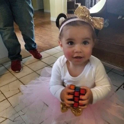 Kendall Bug wearing gold bow headband, wearing a white long-sleeve shirt and pink tutu, holding a partially solved Rubik's Cube.
