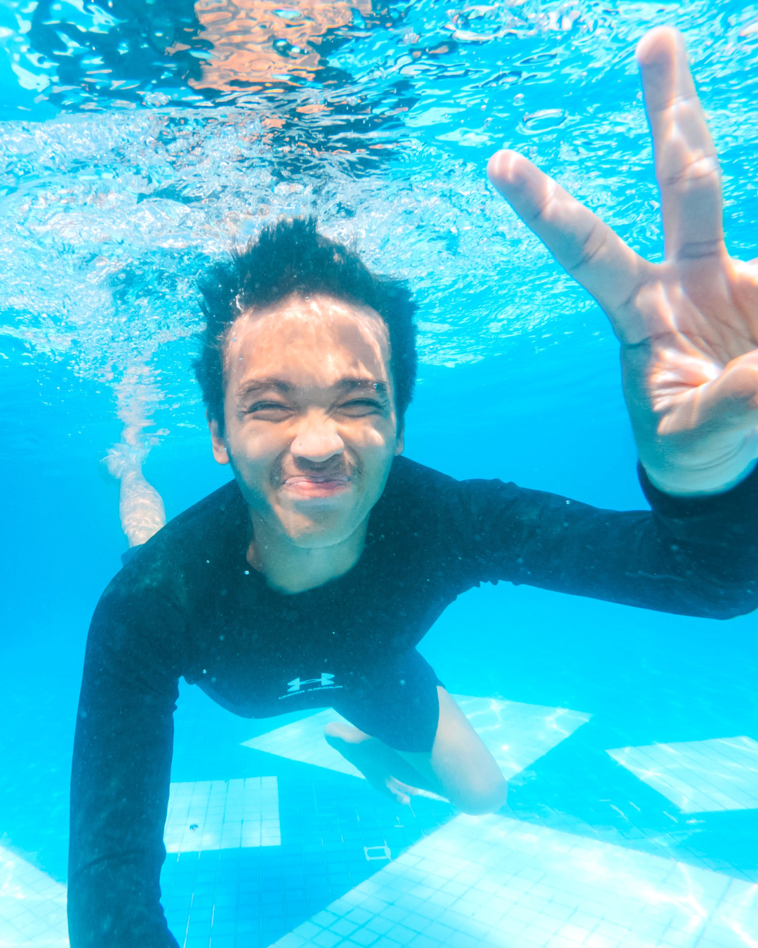 A person underwater making a peace sign with their hand and smiling at the camera.