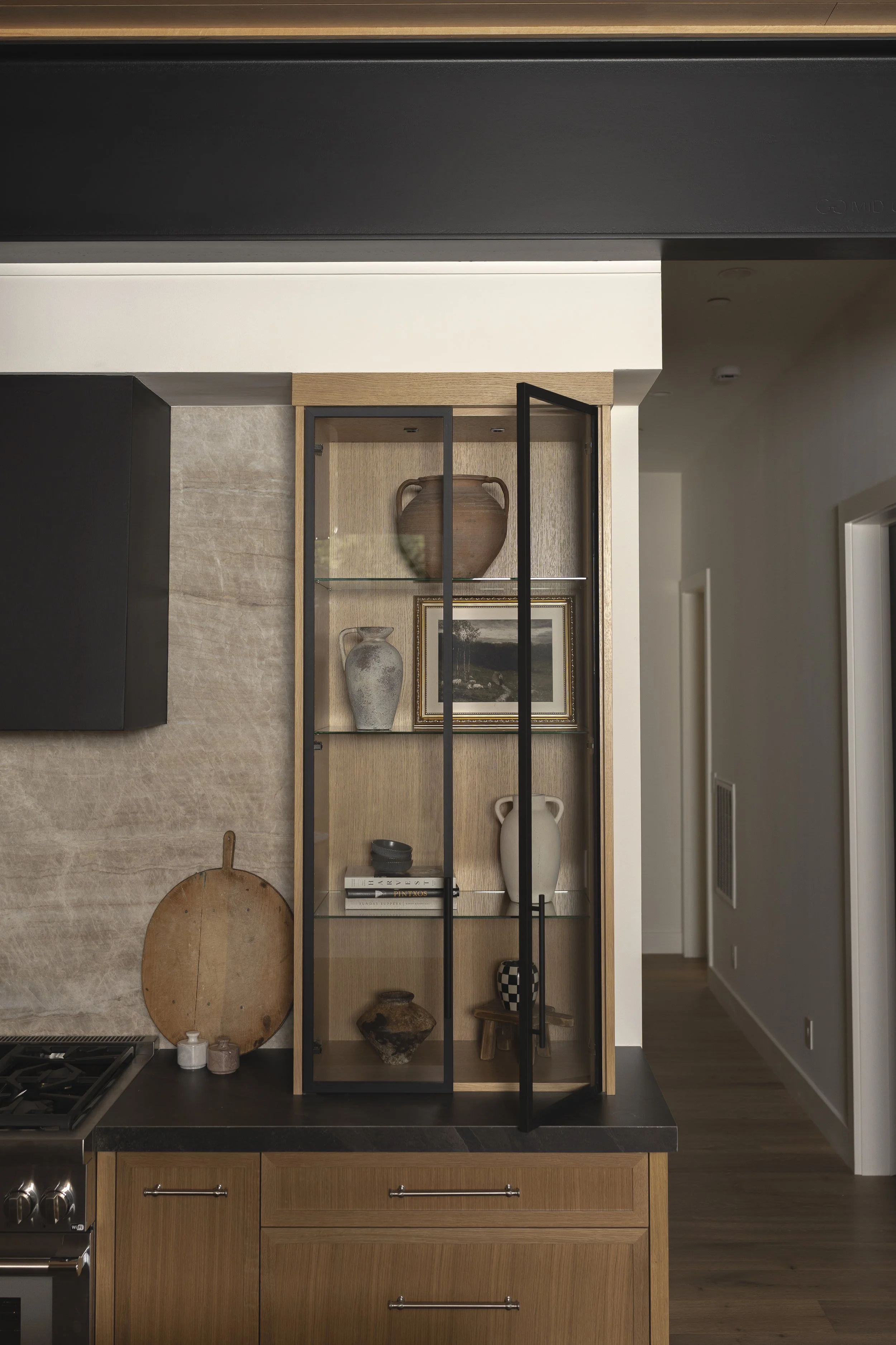 A display cabinet with glass shelves and black metal frame, holding decorative pottery, a framed picture, and books in a modern kitchen.