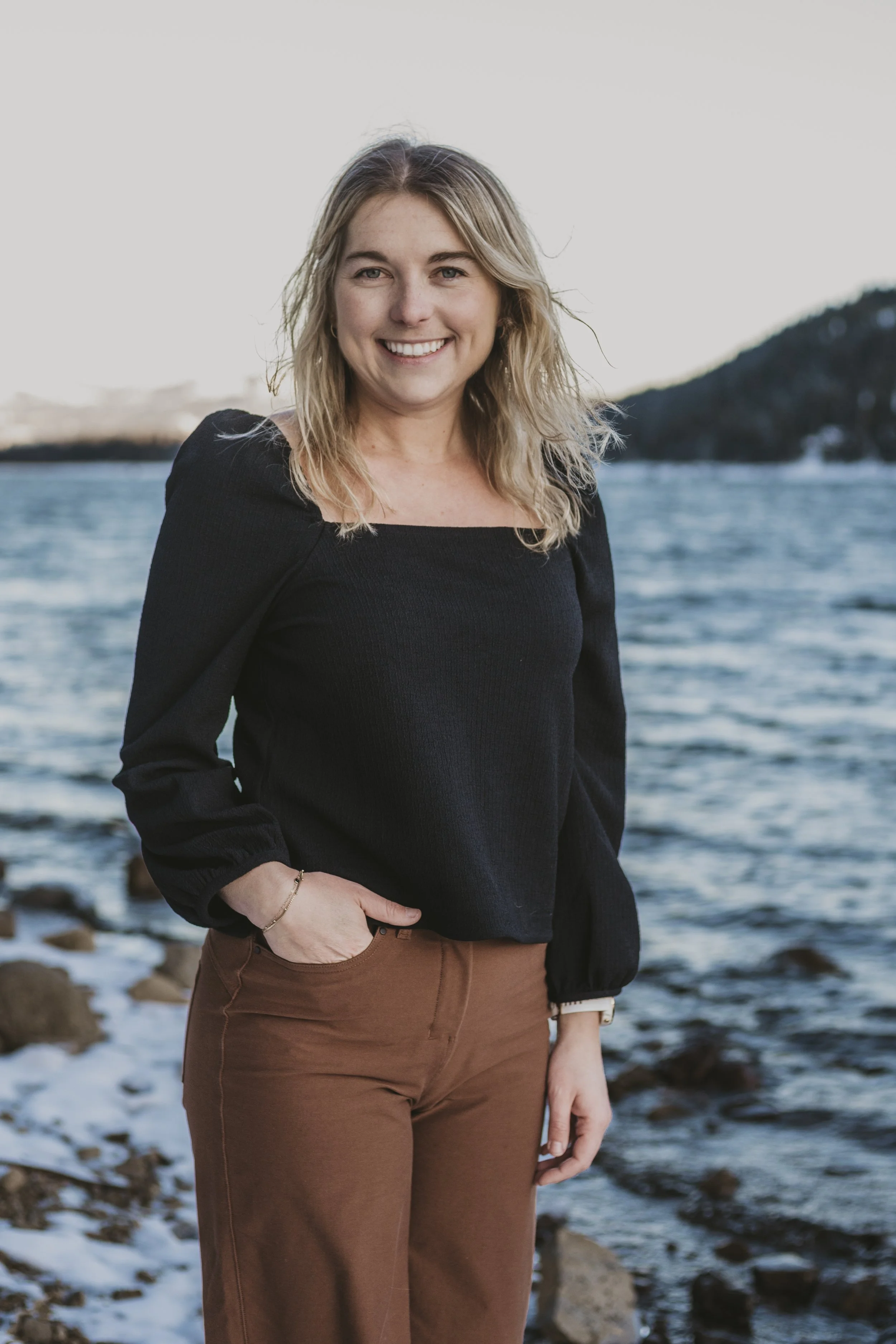 A woman smiling at the camera standing on a rocky shoreline with water and mountains in the background.