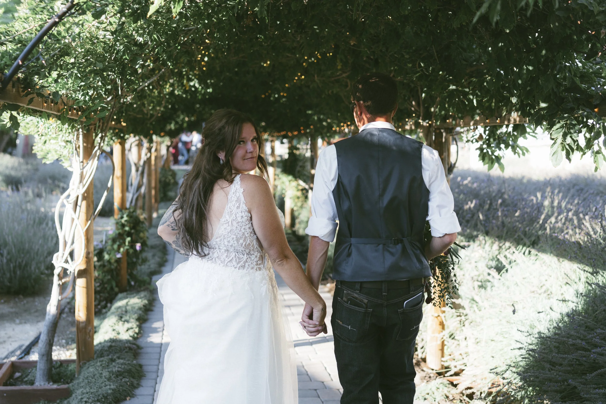 A bride and groom holding hands, walking under a green, leafy archway in a garden setting, with lavender plants in the background.