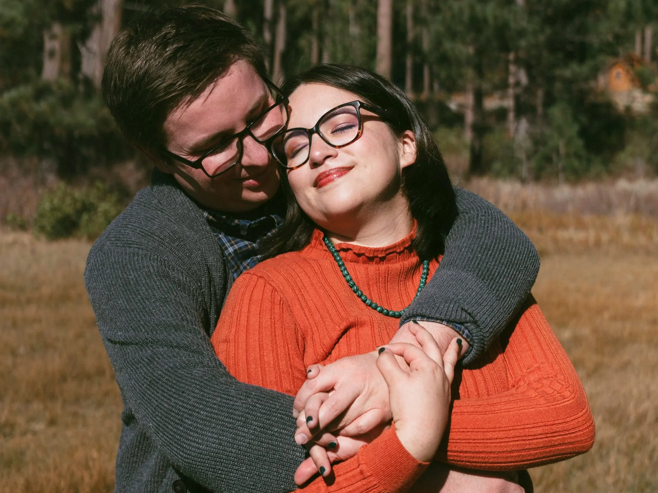 A couple hugging outdoors in a field with trees in the background. The man has dark hair, glasses, and is wearing a grey sweater. The woman has black hair, glasses, and is wearing an orange sweater and a turquoise necklace. Both are smiling with eyes closed.