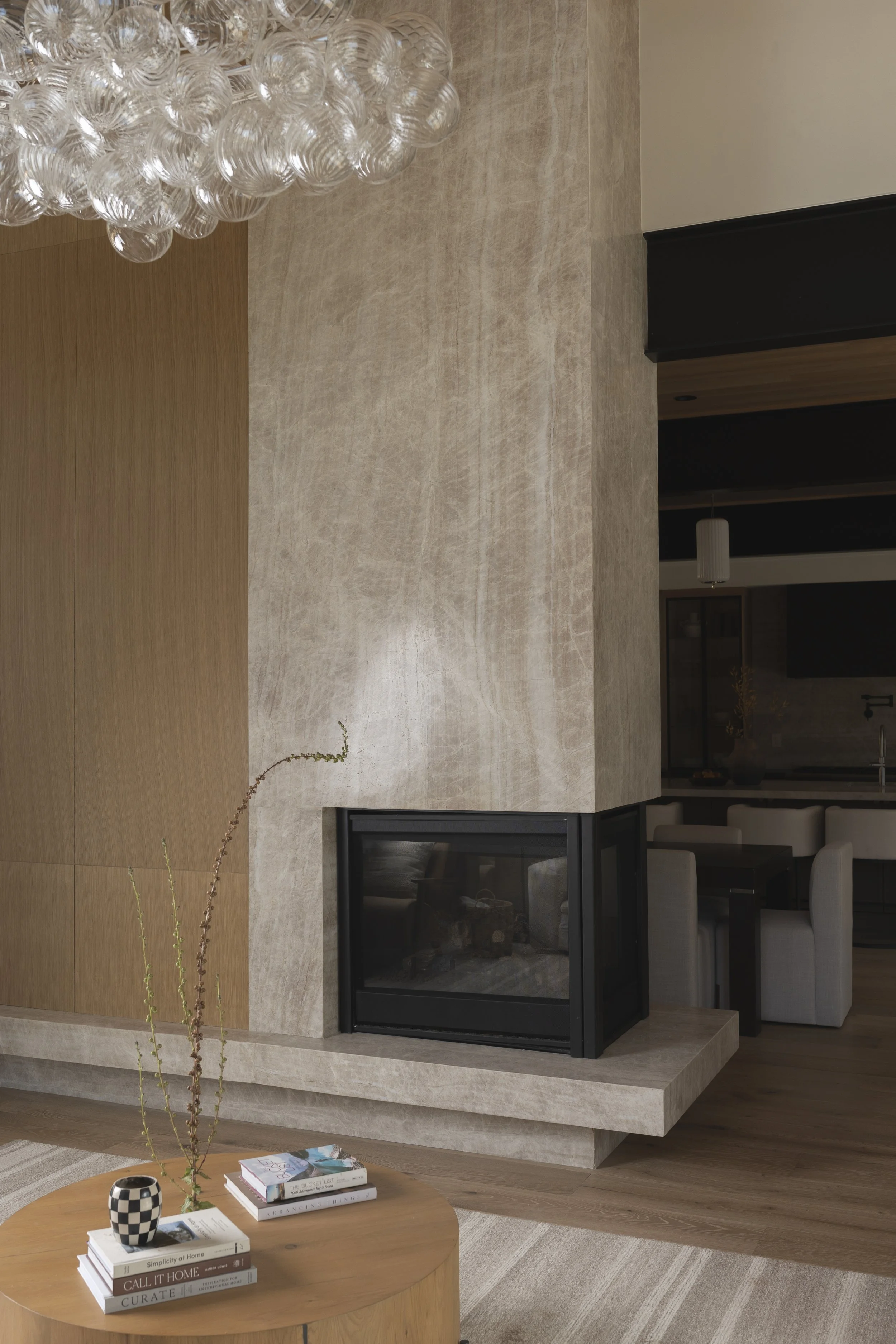 Living room with a modern fireplace, wooden and beige walls, a round wooden coffee table with books and a decorative vase, a glass pendant chandelier, and part of a dining area with a dark table and beige chairs.
