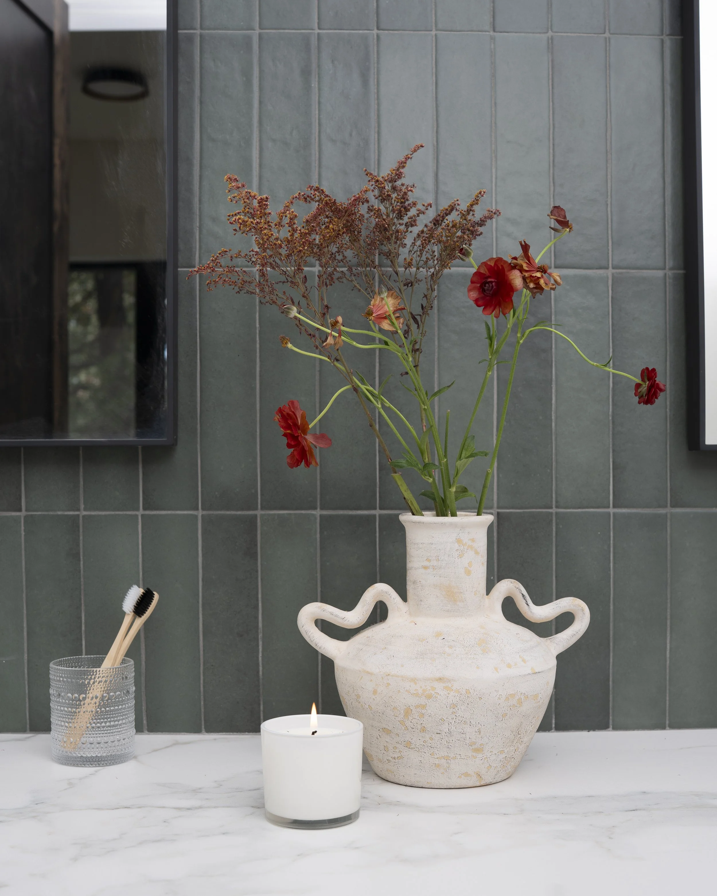 Vase with red and brown flowers on a marble countertop, next to a lit white candle and a container with toothbrushes, set against a green tiled wall.