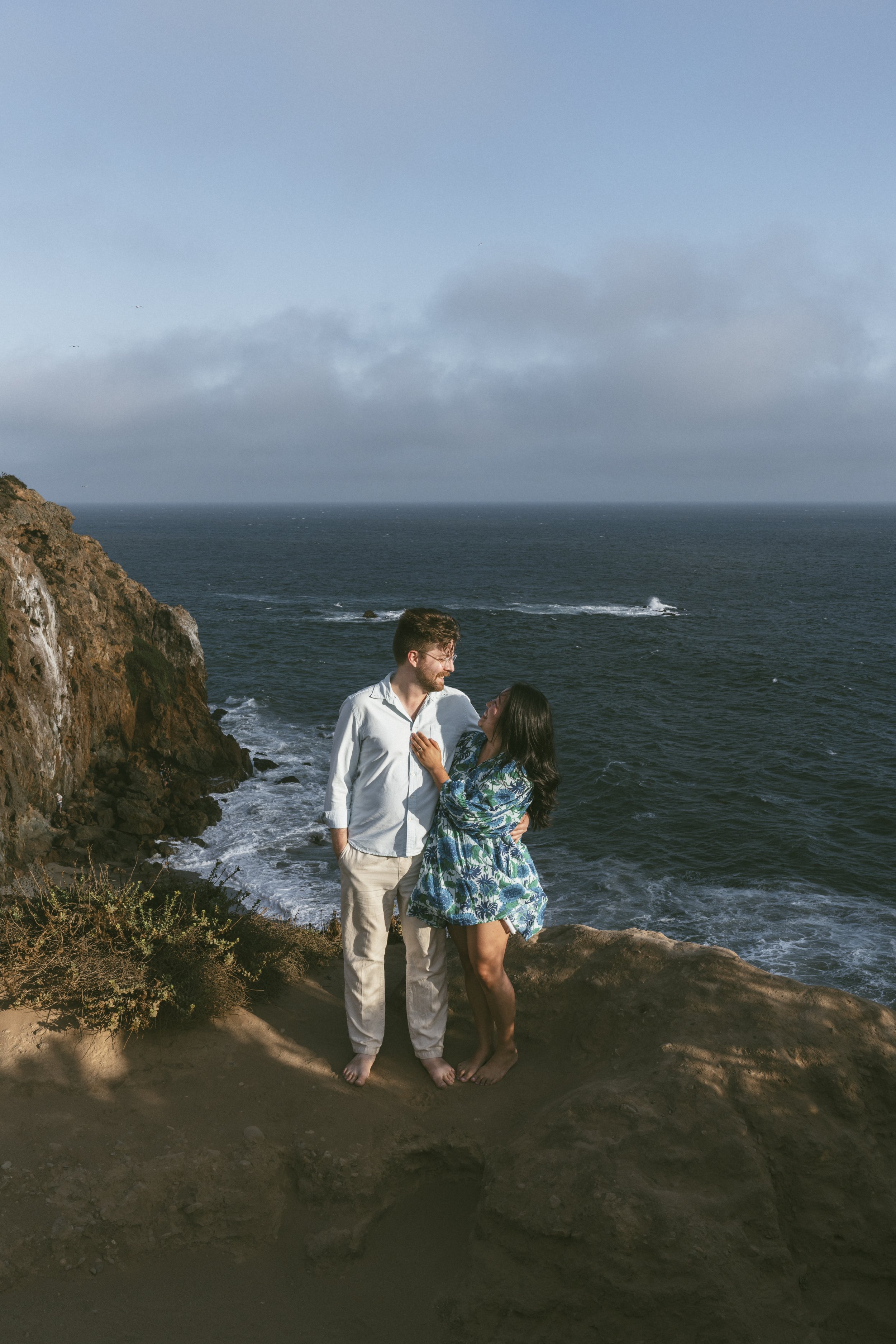 A couple standing on a rocky beach at the cliff edge, overlooking the ocean with cloudy sky in the background. The man has a beard, is wearing a light-colored shirt and pants, and is smiling at the woman. The woman has long black hair, is wearing a blue patterned dress, and is looking up at the man.