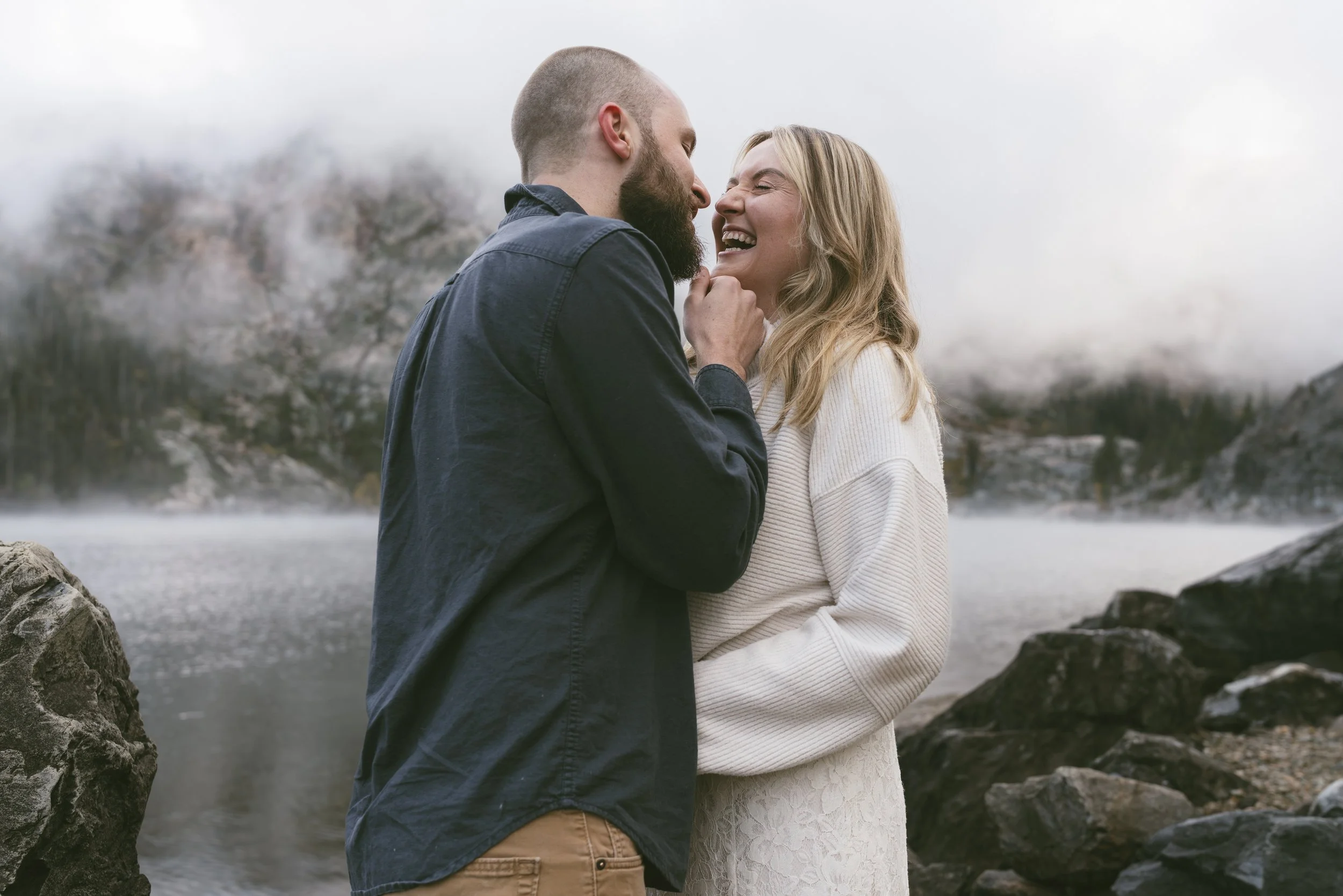A happy couple standing near rocks by a body of water with mountains and mist in the background, enjoying an intimate moment and smiling at each other.