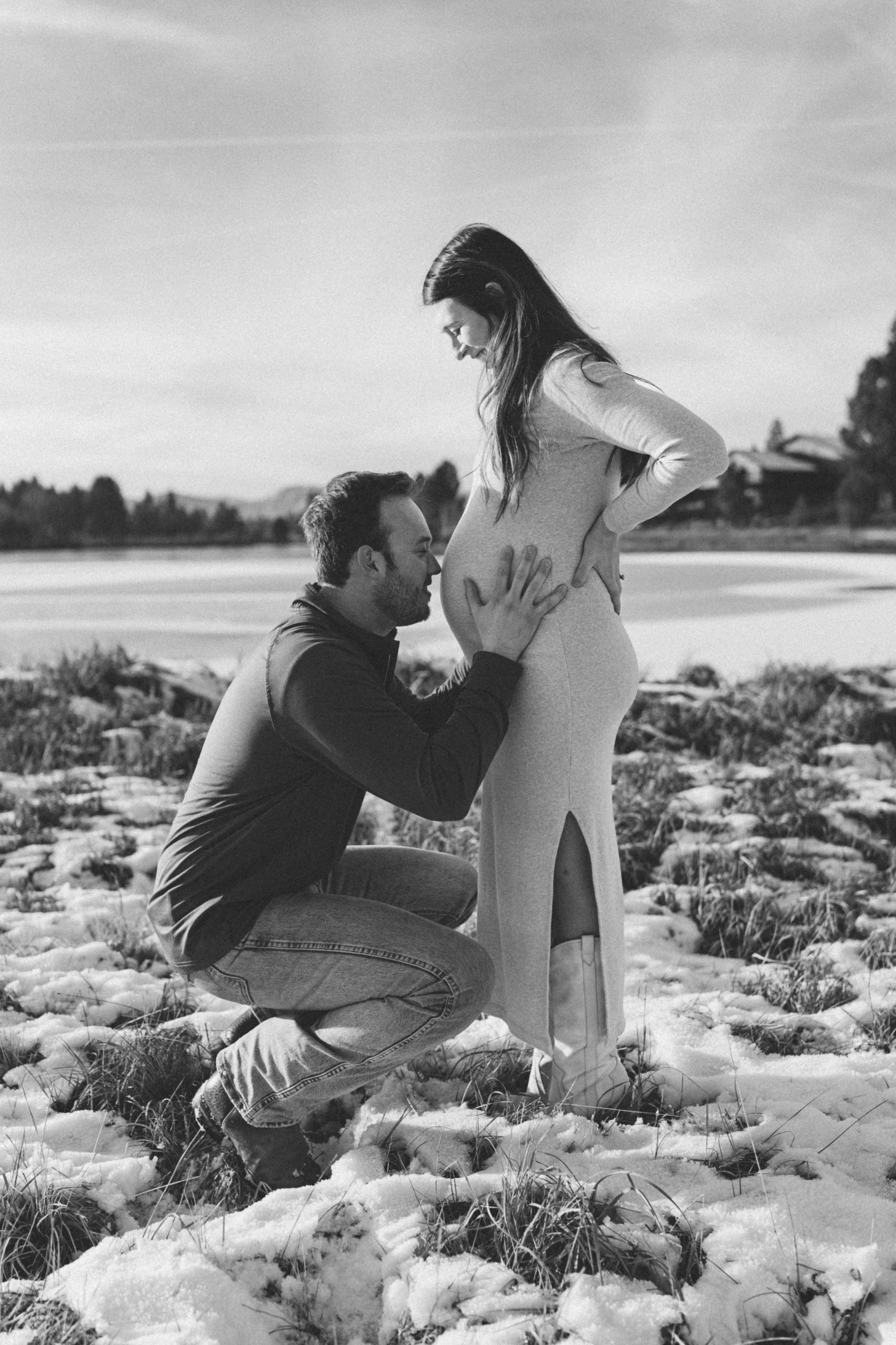 A man kneeling and kissing a pregnant woman on the belly outdoors in a snowy landscape.
