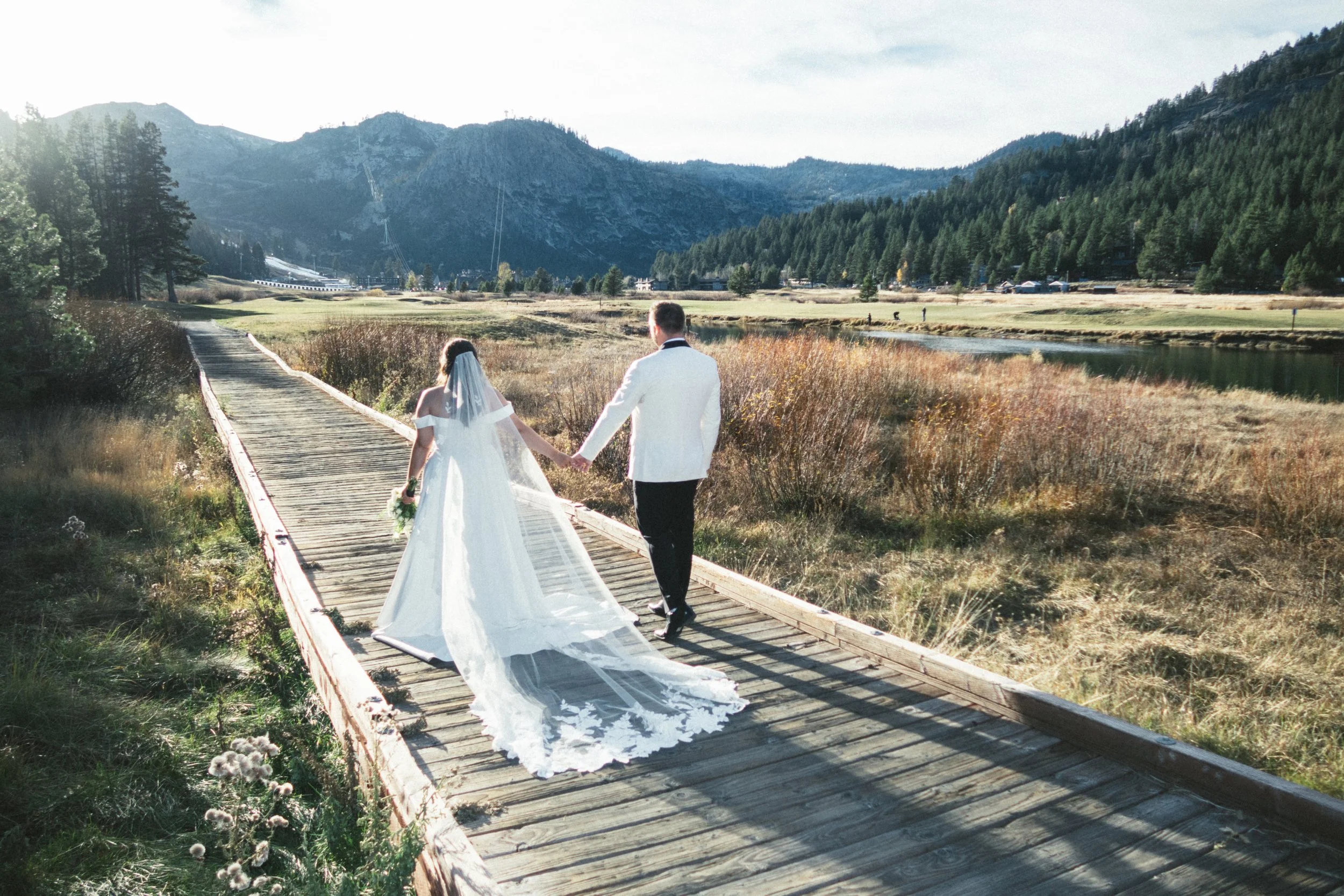 Bride and groom walking hand in hand on a wooden bridge in a scenic outdoor setting with mountains, trees, fields, and a lake.