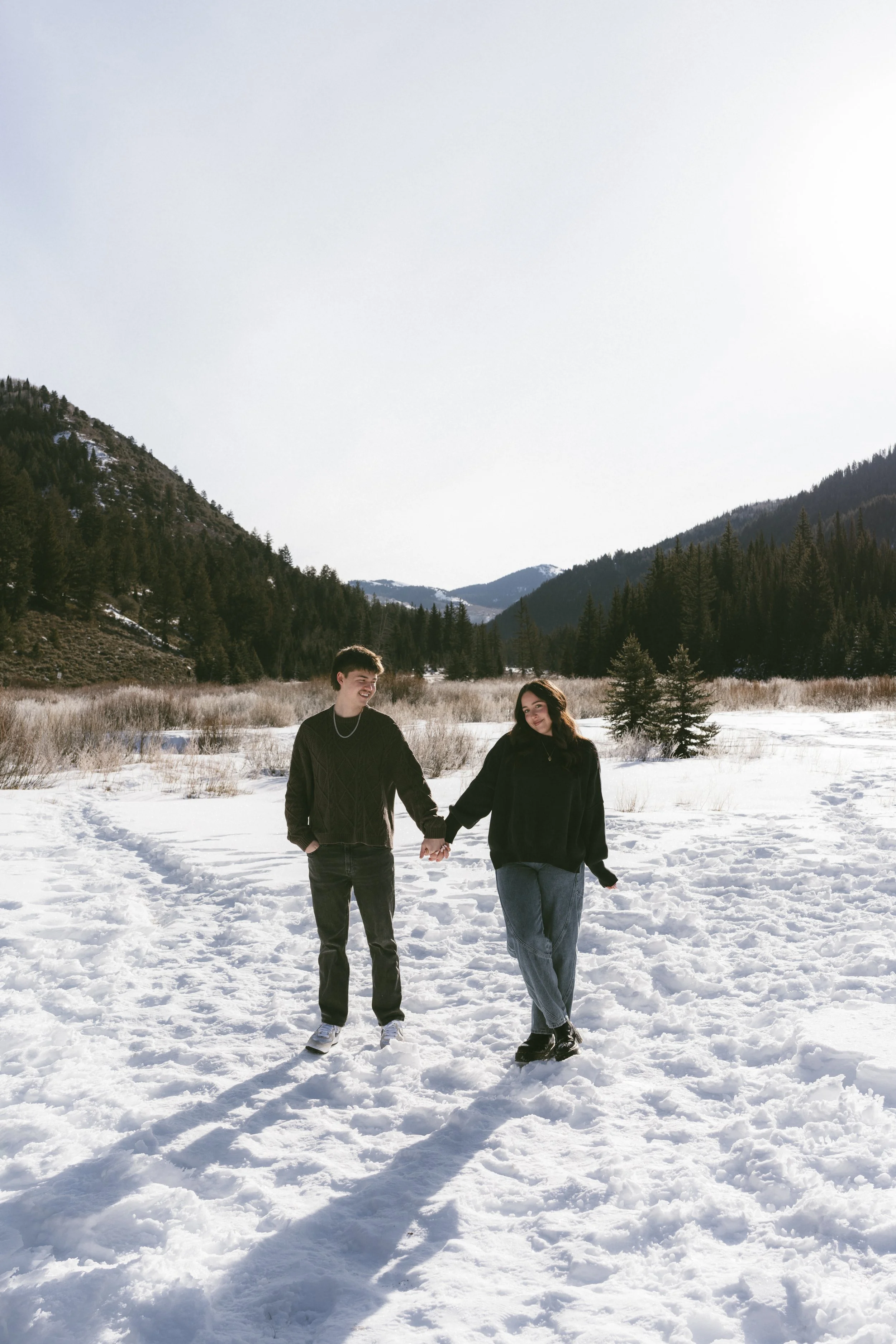 A young man and woman holding hands walk through snow in a mountain valley with evergreen trees and mountains in the background.