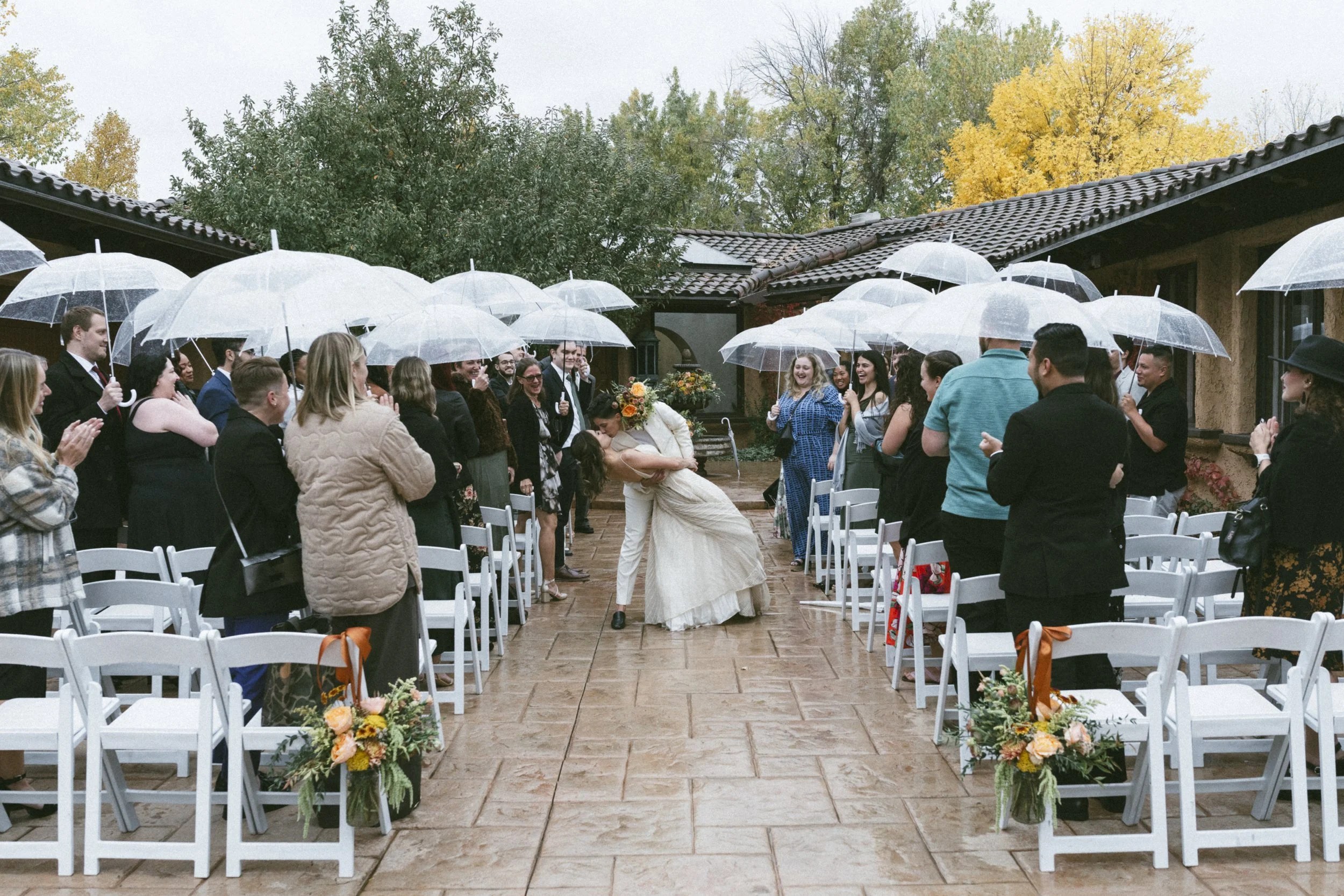 Bride and groom kiss as guests hold umbrellas during outdoor wedding ceremony on a rainy day, with autumn trees in the background.