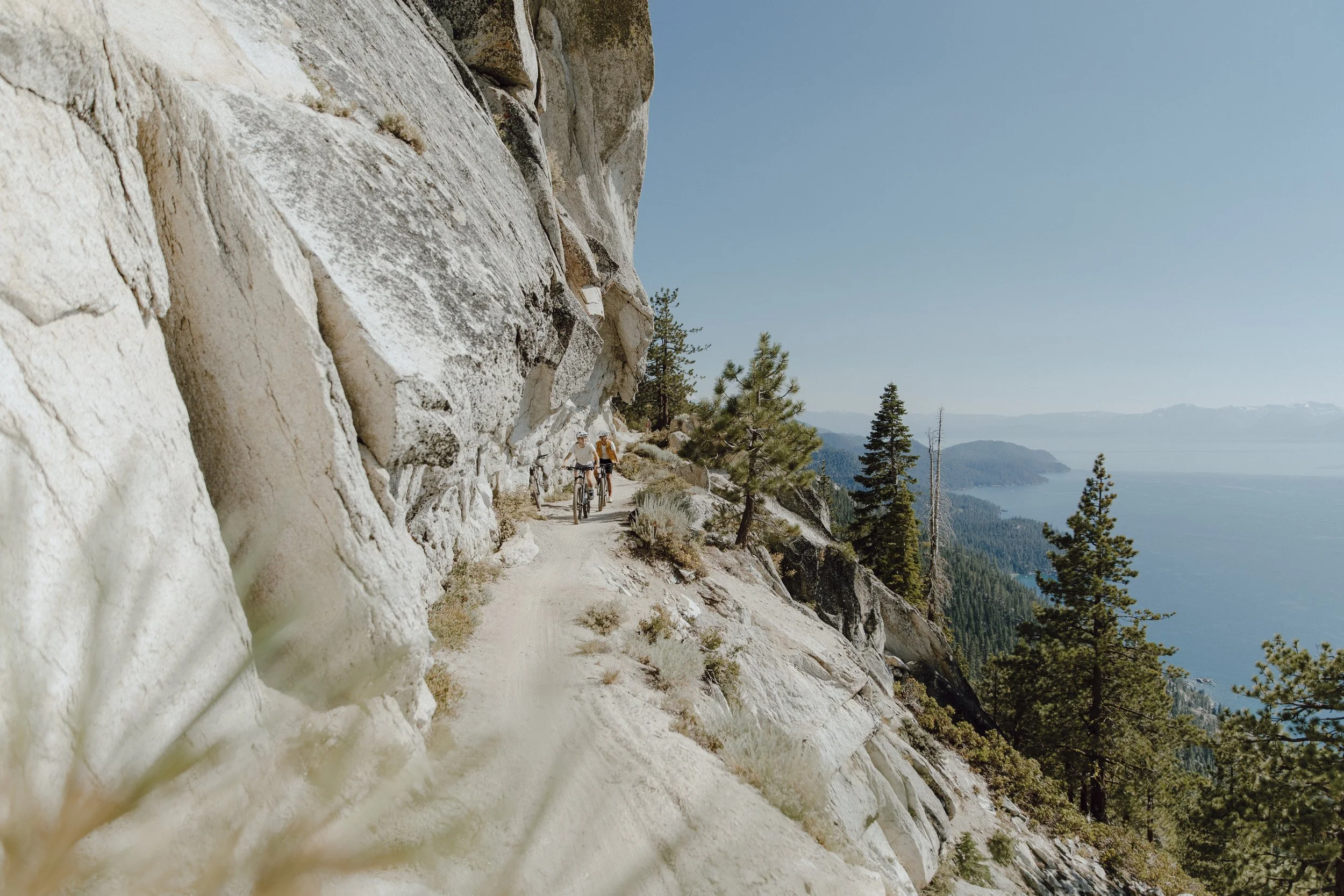 Two people riding mountain bikes on a narrow dirt trail alongside a rocky cliff with pine trees, overlooking a lake and distant mountains.