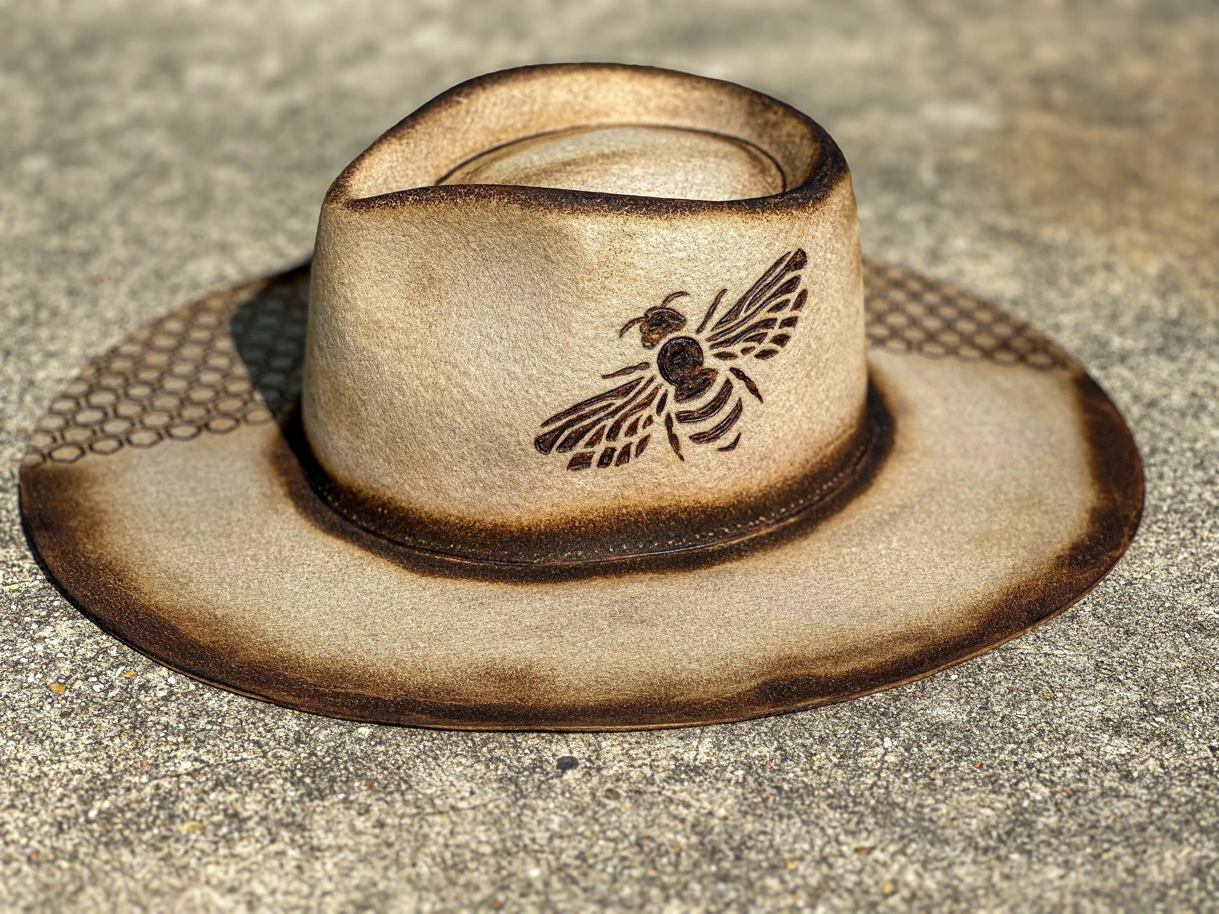 A beige cowboy hat with a black bee design on the front, resting on a textured surface.