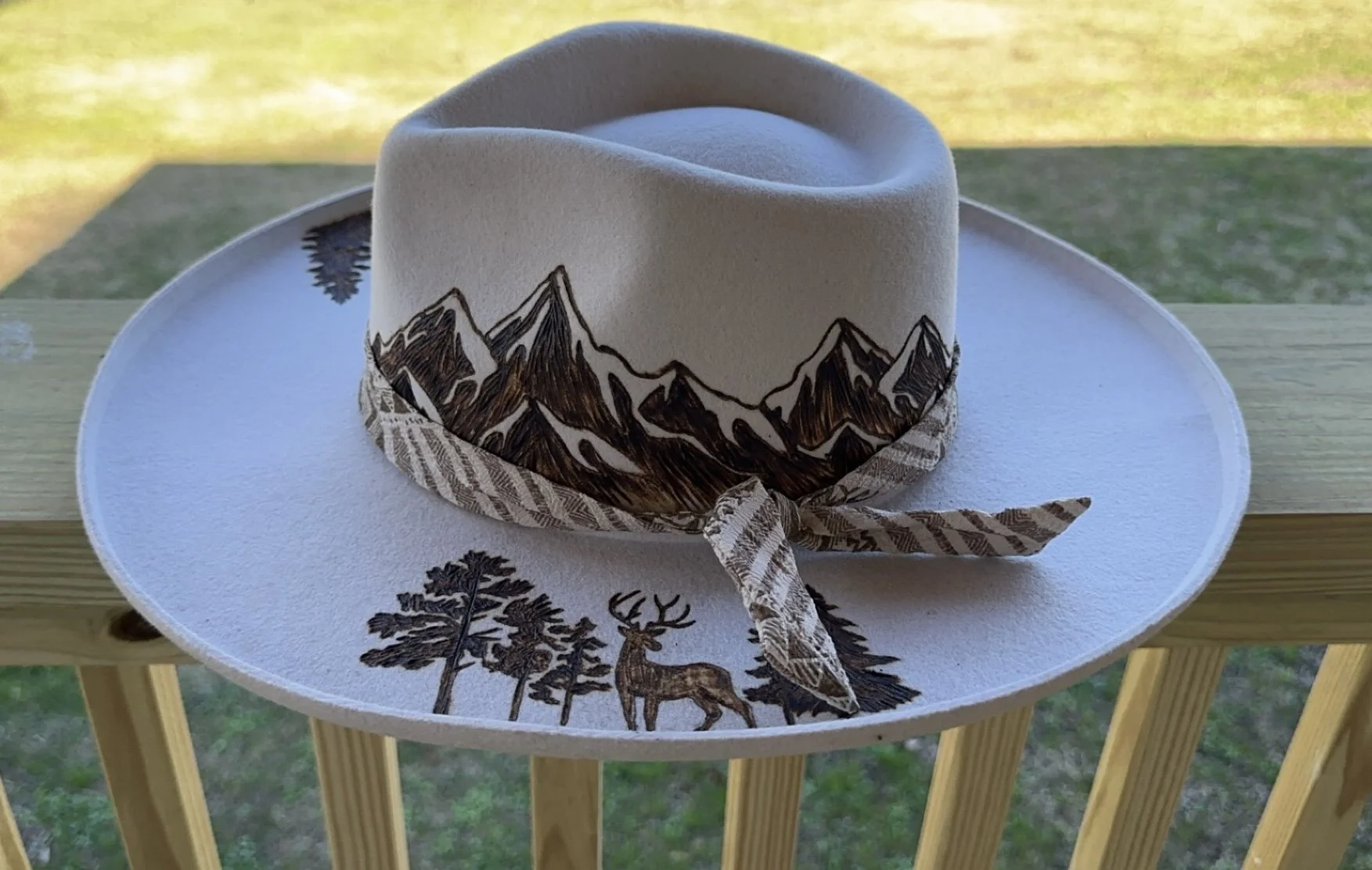 A white cowboy hat with mountain, tree, and deer designs on the band and brim, sitting on a white platter on a wooden railing, with a grassy field in the background.