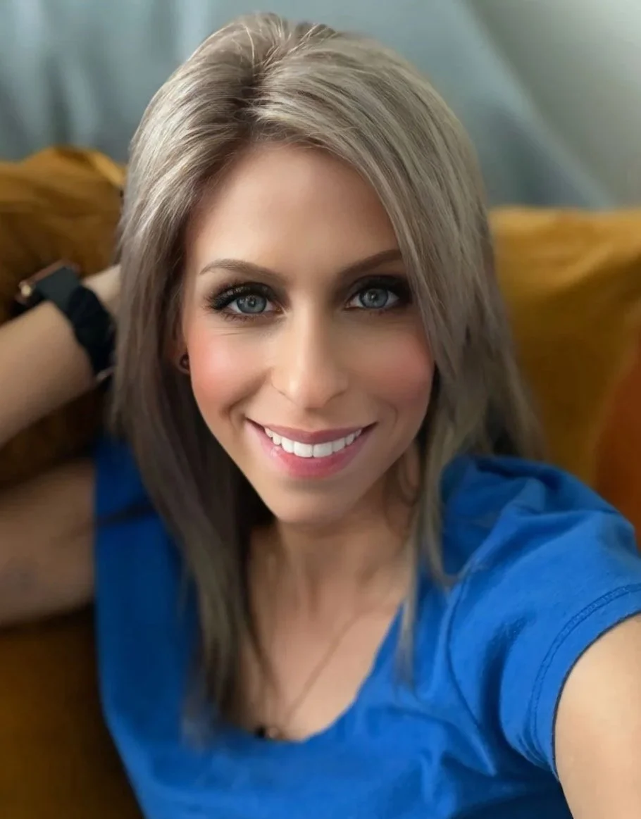 A woman with blonde hair and blue eyes smiling at the camera, wearing a blue shirt, sitting on a yellow couch or pillow.
