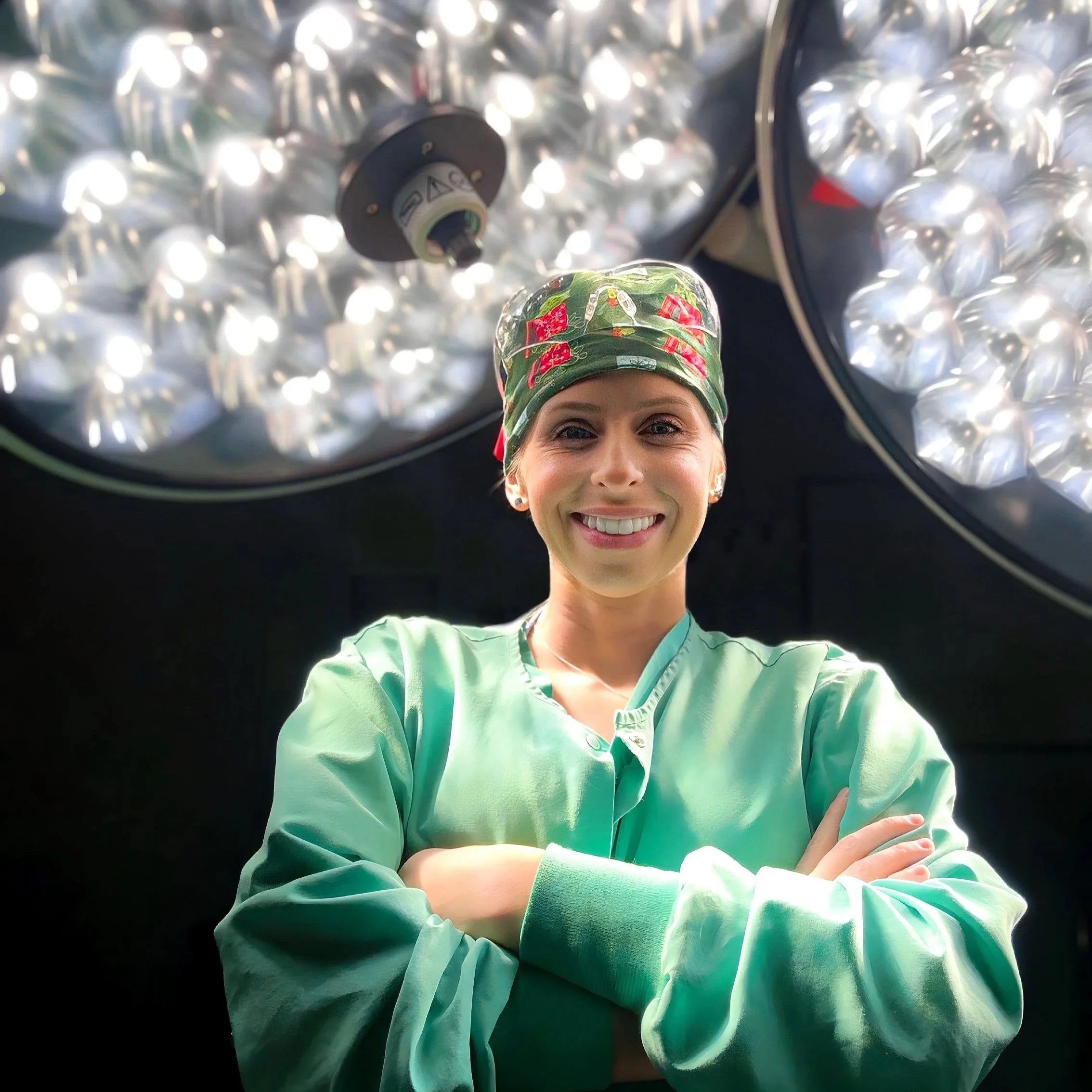 Female surgeon in scrubs and cap standing in an operating room with two large surgical lights above.