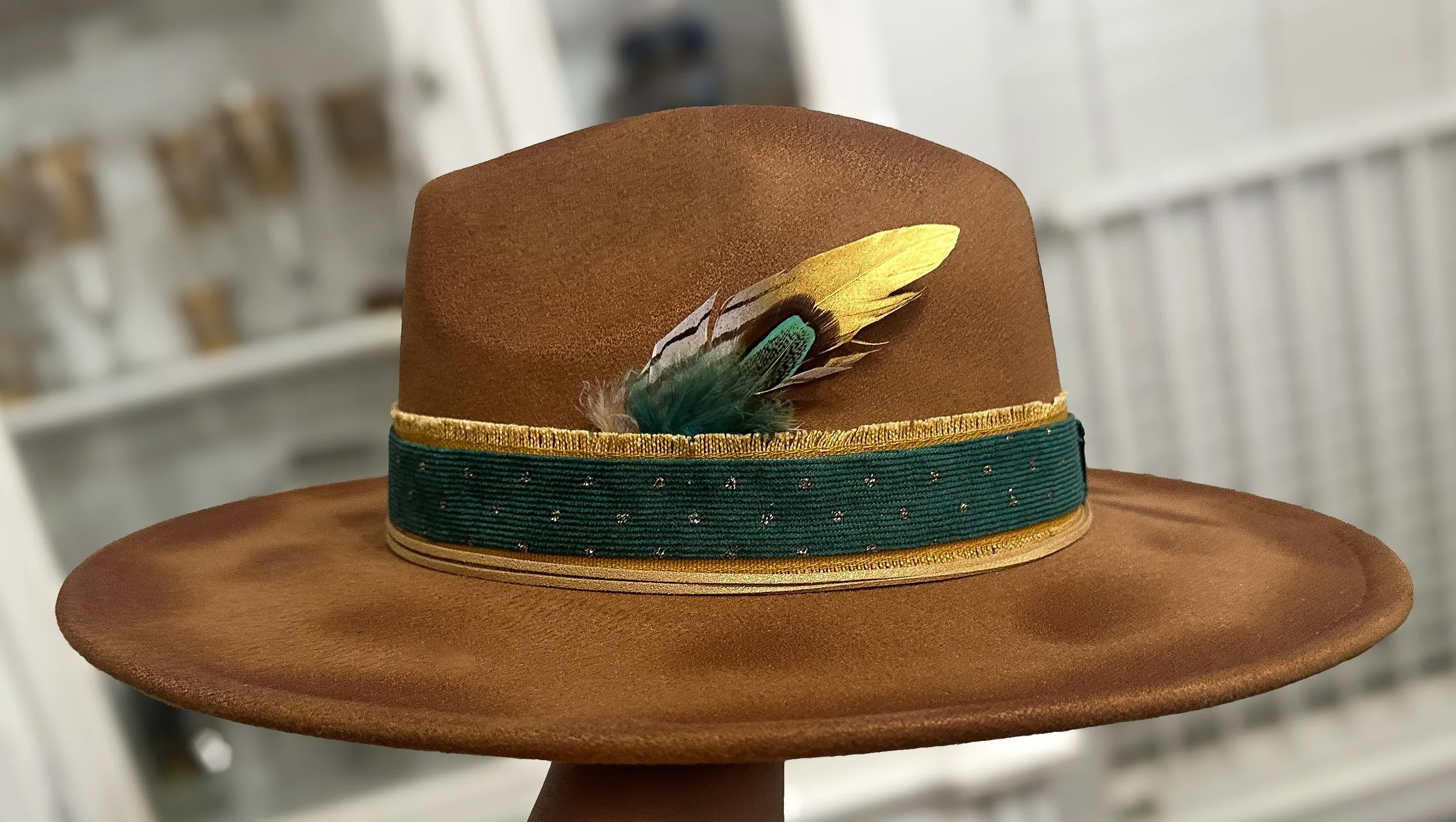 Brown felt hat with decorative gold trim, a dark green band with small gold dots, and a multicolored feather on the side.