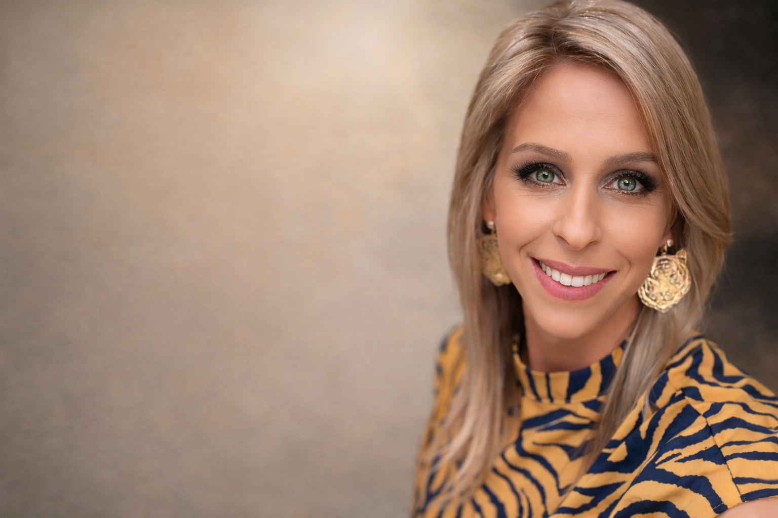 A woman with blonde hair, blue eyes, wearing a yellow and navy zebra print top, large gold earrings, and smiling