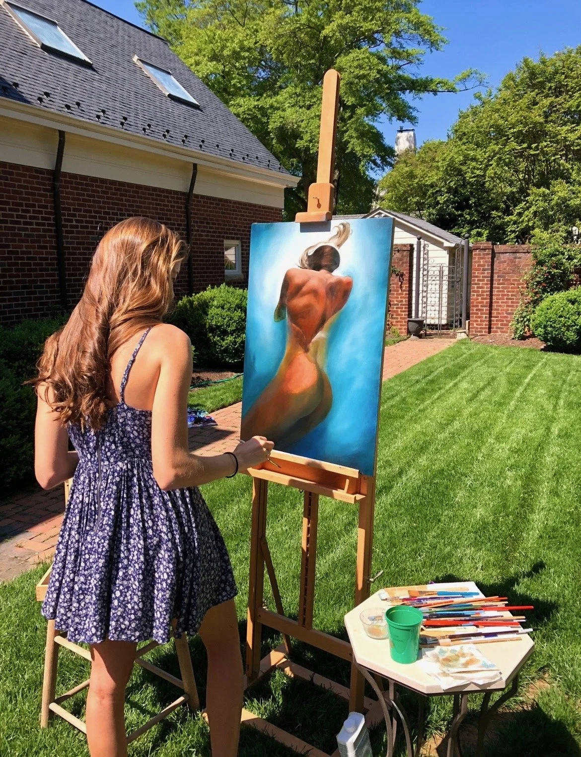 A young woman with long, wavy, reddish-brown hair, wearing a blue floral dress, is painting outdoors on an easel. The painting depicts a rear view of a nude woman with her back turned, set against a bright blue background. There are painting supplies on a nearby table, and the scene is in a sunny, green garden with a brick house and trees in the background.