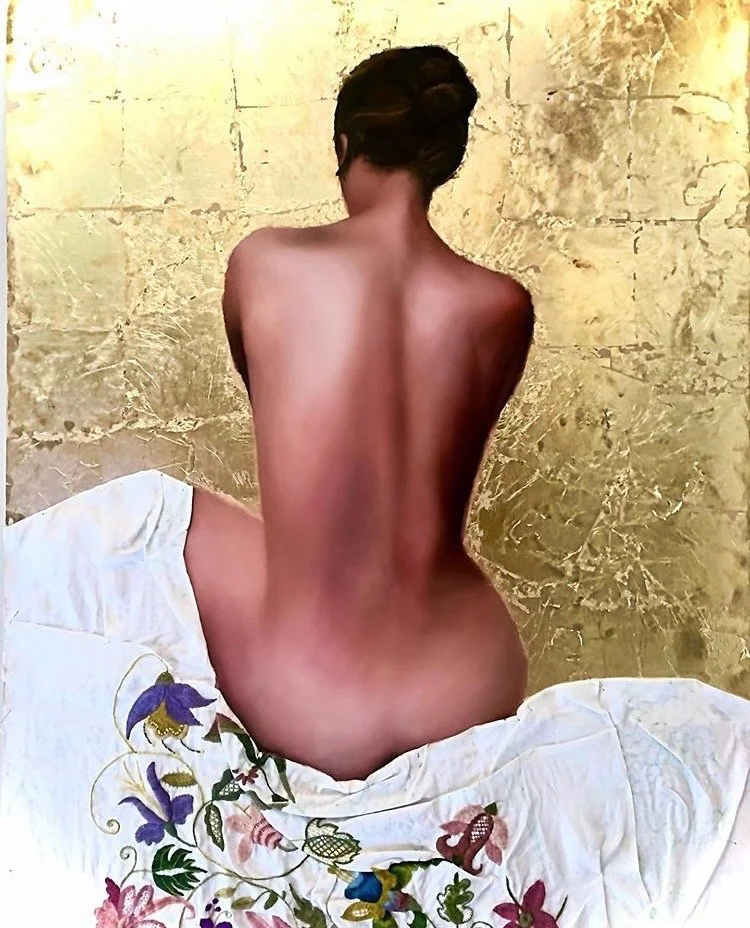 A woman with short dark hair seated topless on a bed with colorful floral embroidery, facing away from the camera, with a gold textured wall in the background.