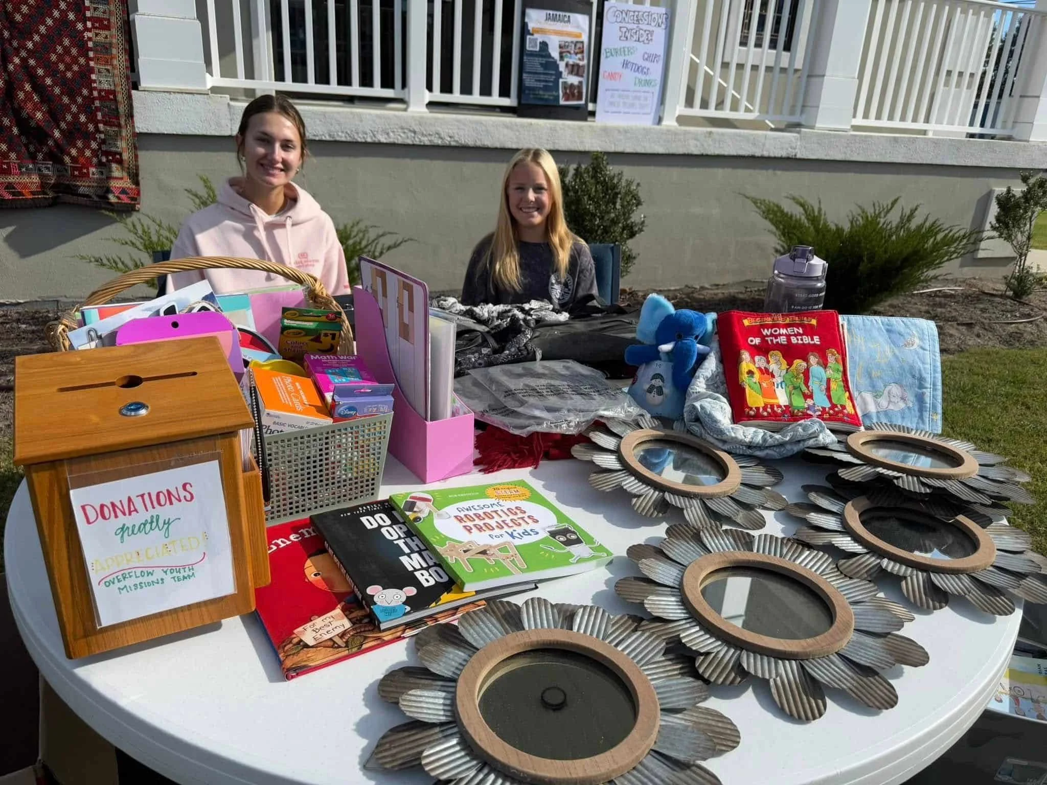 Two smiling women sit behind a table with books, toys, mirrors, and a donation box at an outdoor fundraiser event.