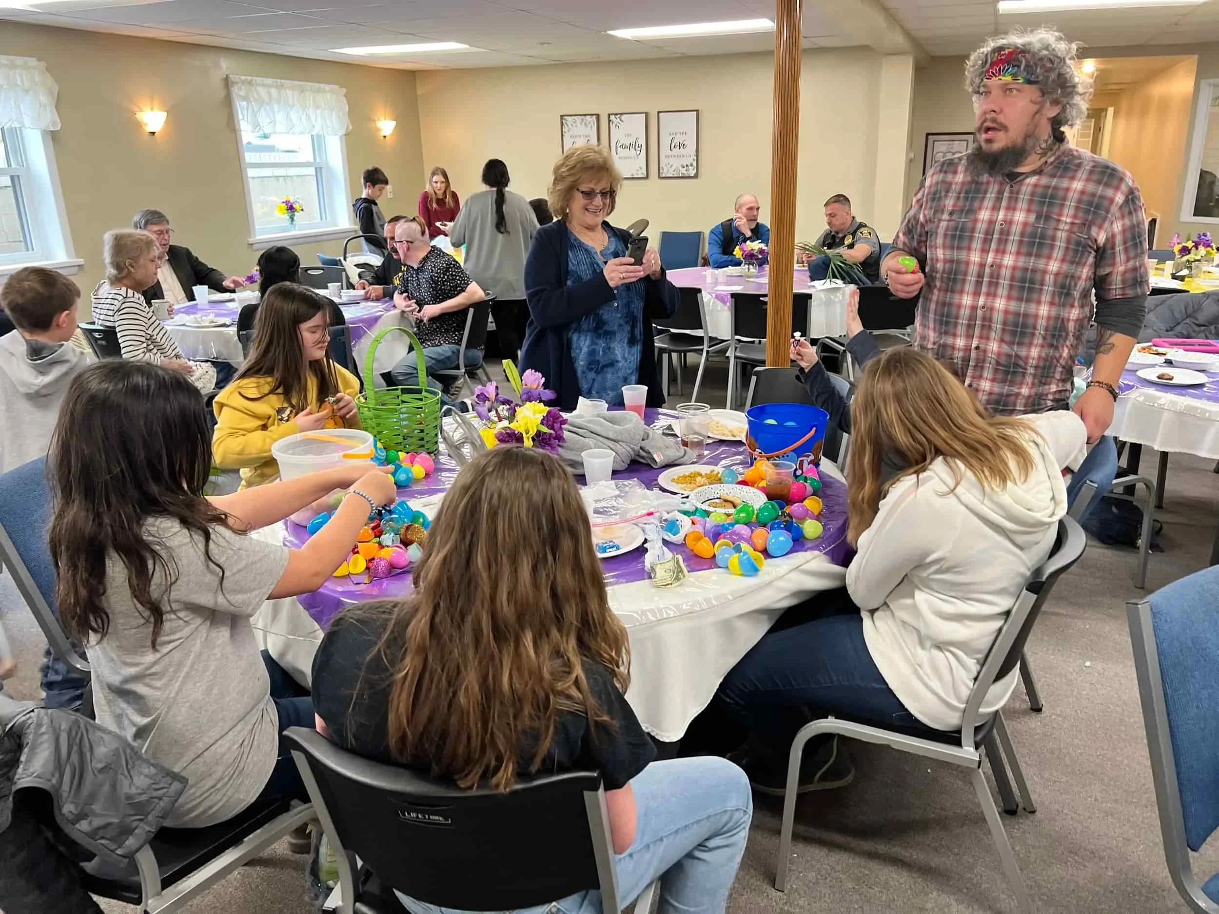 People gather around tables decorating colorful Easter eggs in a bright, festive room with spring decorations.