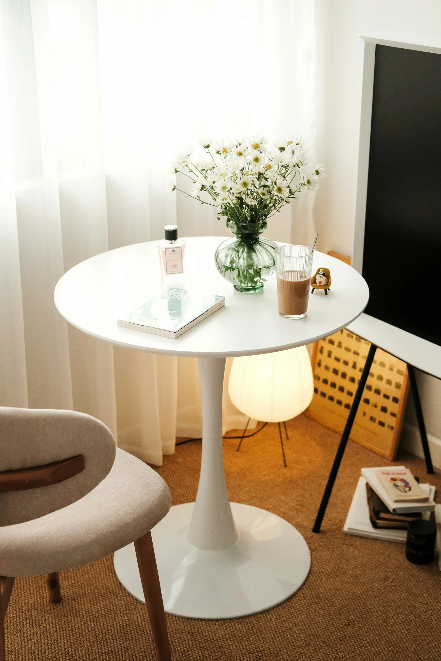A round white table with a glass vase of white daisies, a bottle of perfume, a book, and a glass of chocolate milk. Nearby are a beige chair, a white lamp on the floor, a black TV, and a stack of books.