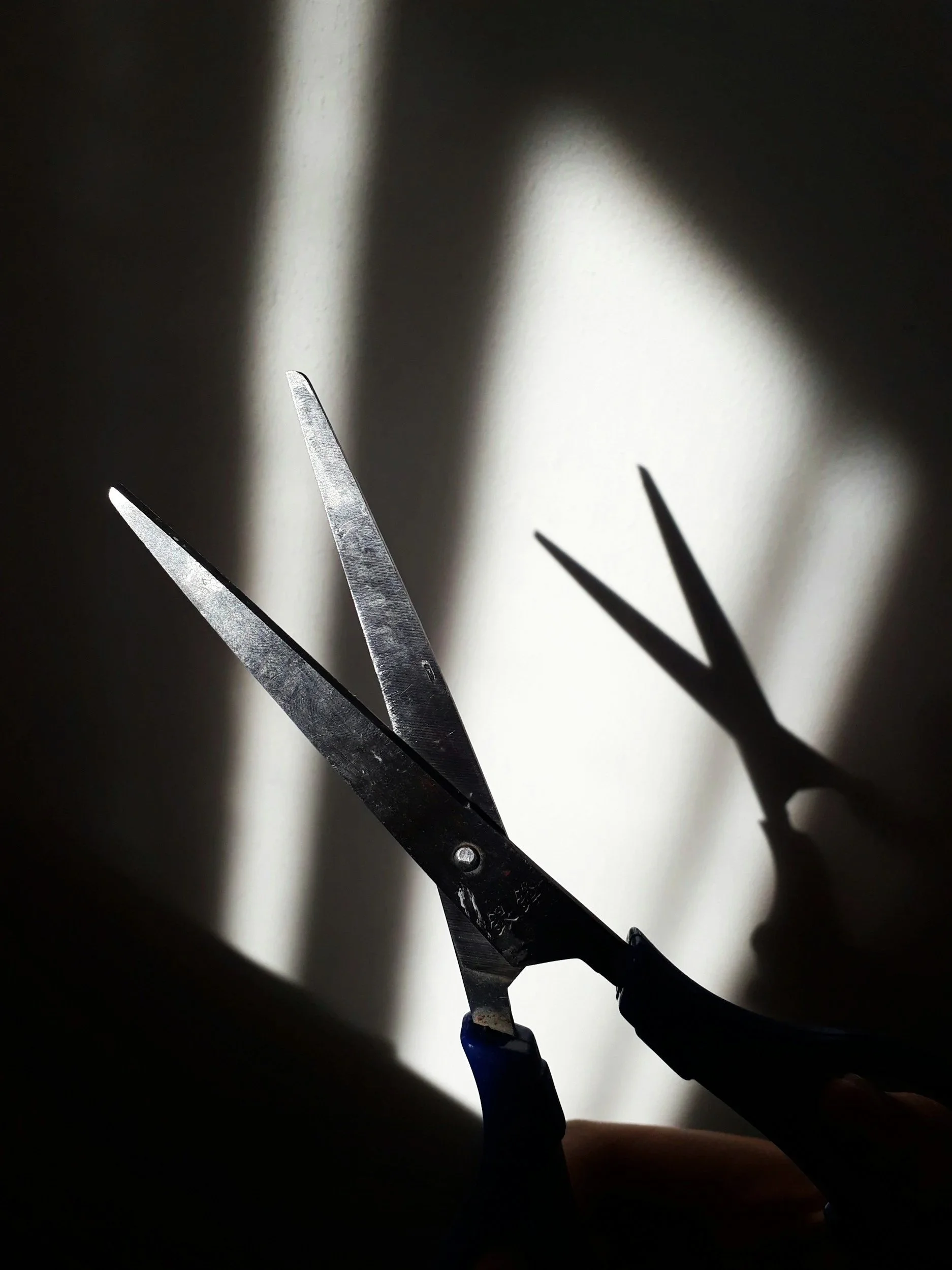 A pair of scissors held against a wall with shadows cast by sunlight, creating a shadow of another pair of scissors.