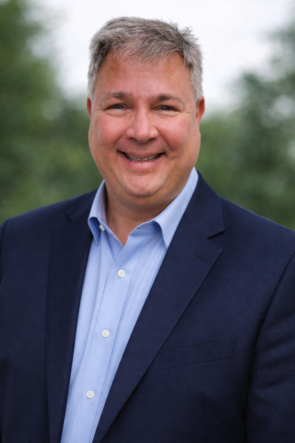 A smiling man in a navy blazer and light blue shirt standing outdoors with green trees in the background.