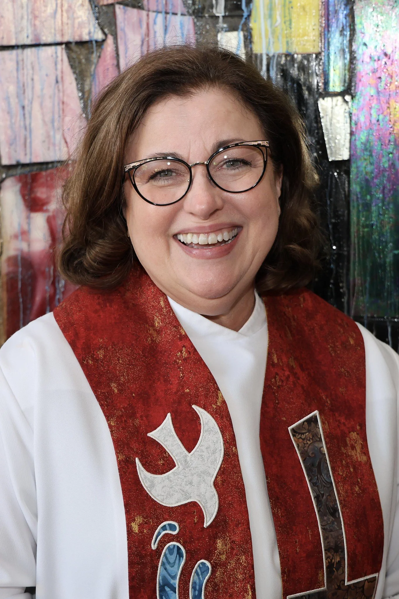 A photo of Pastor Anna: a woman with glasses smiling, wearing a white robe with a red patterned stole that has a dove and cross embroidered on it, standing in front of a colorful abstract background.
