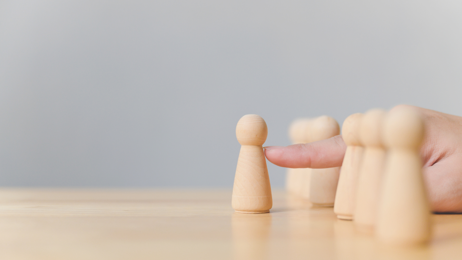 A person gestures to a single wooden figurine while four other figurines stand in a row on a wooden surface.